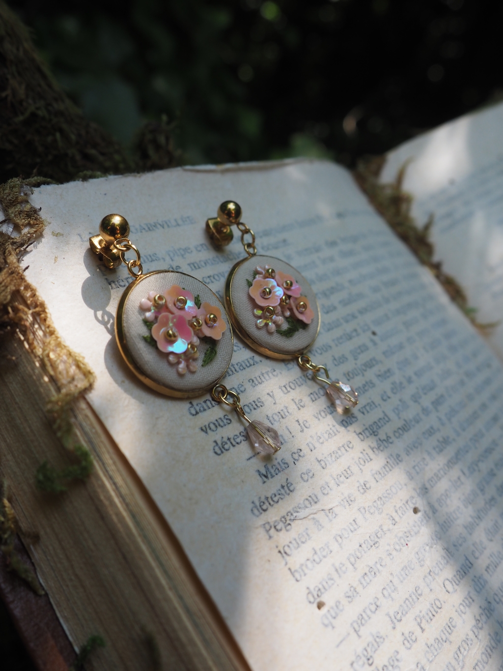 Cette image montre une paire de boucles d'oreilles délicates posées sur un livre ouvert. Les boucles d'oreilles semblent être de style vintage ou romantique, avec des médaillons ronds ornés de petites fleurs roses et de détails décoratifs. Elles sont montées sur ce qui semble être un fond nacré ou blanc, avec des petites breloques qui pendent au bas. Les boucles d'oreilles reposent sur les pages d'un livre avec du texte imprimé, créant une composition très poétique et romantique. La photo est prise avec une belle profondeur de champ qui rend l'arrière-plan légèrement flou, mettant en valeur les bijoux au premier plan.