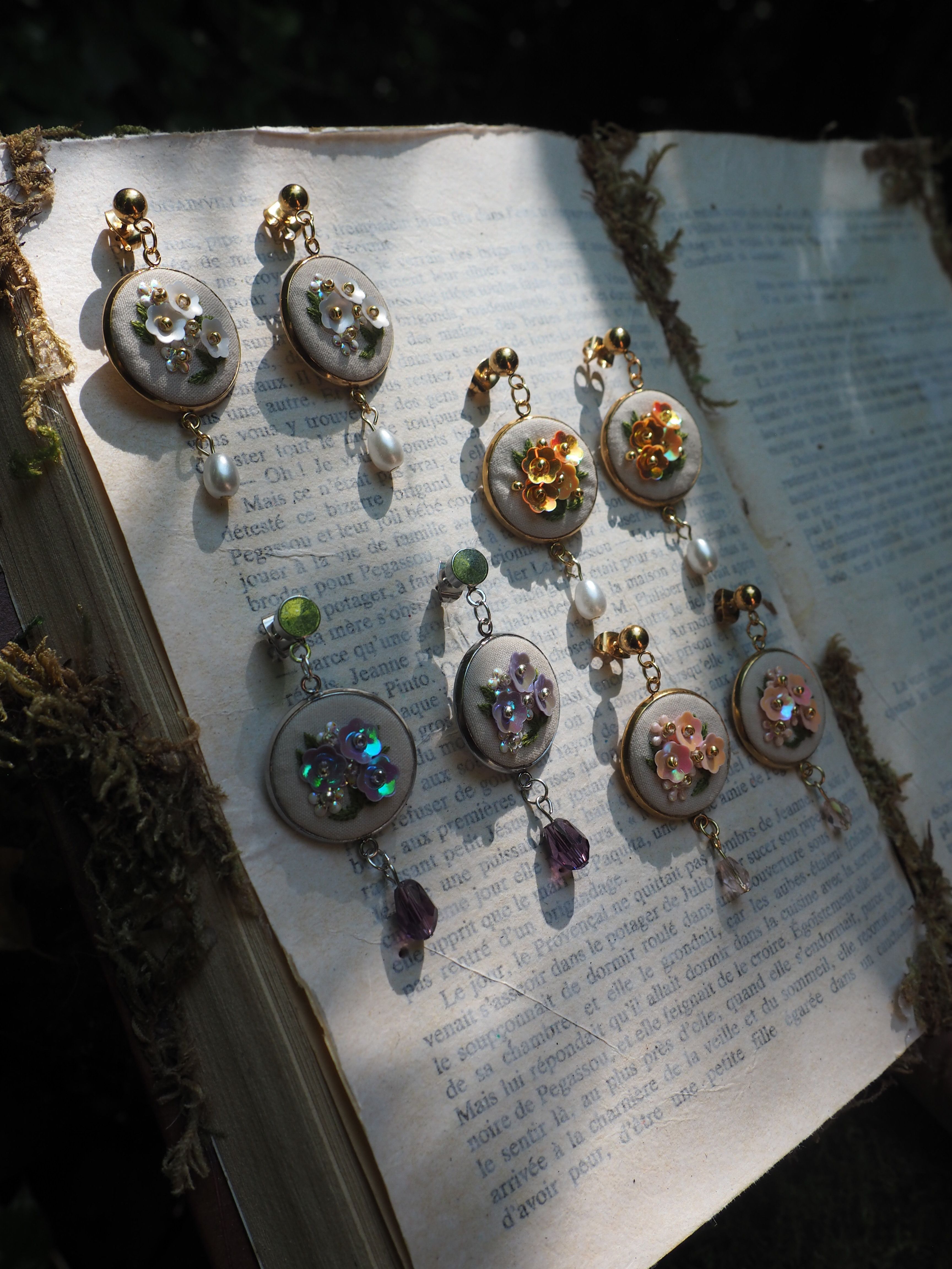 L'image montre une collection de petits bijoux ou ornements disposés sur ce qui semble être des pages d'un livre ou d'un document écrit. Ces ornements paraissent être des boutons ou des pendentifs décoratifs, certains avec des pierres ou des cristaux de différentes couleurs (on peut distinguer des teintes dorées, vertes, bleues et violettes). Les pièces semblent avoir une base blanche ou argentée et sont arrangées de manière ordonnée sur le texte. La lumière crée des ombres intéressantes sur les pages, donnant à l'image une qualité artistique et délicate. C'est une composition qui évoque un peu l'artisanat ou la bijouterie artisanale.