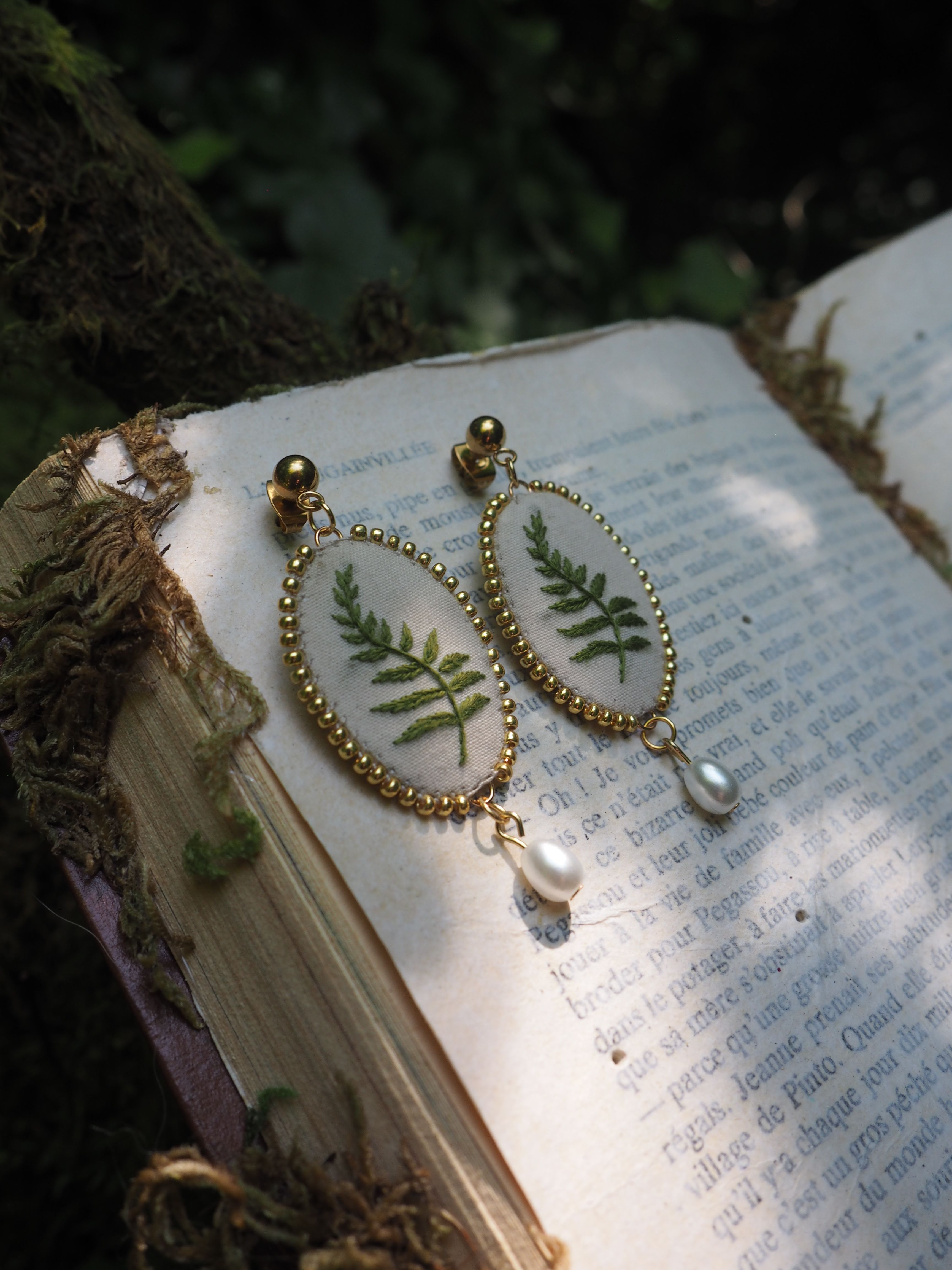 Cette image montre une paire de boucles d'oreilles élégantes et délicates posées sur un livre ouvert dans ce qui semble être un cadre naturel en extérieur. Les boucles d'oreilles sont de style vintage avec des contours dorés en perles et contiennent chacune une véritable fougère pressée et séchée à l'intérieur, encapsulée sous verre. Au bas de chaque boucle d'oreille pend une petite perle ou pierre transparente. Le design est très romantique et naturel. Les boucles d'oreilles reposent sur les pages d'un livre ancien ou vintage, dont on peut apercevoir le texte imprimé en arrière-plan. L'image a une ambiance très poétique et bucolique, avec un éclairage naturel doux qui met en valeur les détails délicats des bijoux.