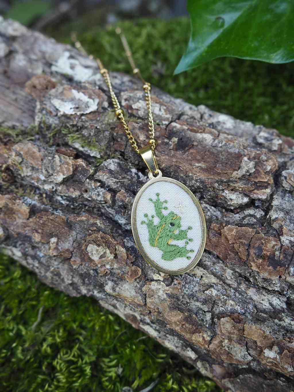 L'image présente un collier avec un pendentif ovale, posé sur un morceau d'écorce d'arbre. Le pendentif est de couleur dorée et contient une broderie d'une grenouille verte sur un fond clair, probablement blanc ou crème. La grenouille est représentée de manière stylisée, avec des détails de broderie qui lui donnent du relief. Le collier est une chaîne fine et dorée, qui s'étend au-dessus et en dessous du pendentif, suggérant qu'il est posé sur l'écorce. L'écorce est rugueuse et texturée, avec des tons de brun et de gris, et est recouverte par endroits de mousse verte, ce qui crée un contraste intéressant avec le pendentif. En arrière-plan, on peut distinguer des éléments de verdure flous, notamment une feuille verte brillante en haut à droite, ce qui renforce l'ambiance naturelle et forestière de la scène. La lumière semble naturelle et douce, mettant en valeur les couleurs et les textures. L'ensemble de l'image dégage une atmosphère calme et organique, soulignant le caractère artisanal et naturel du bijou.