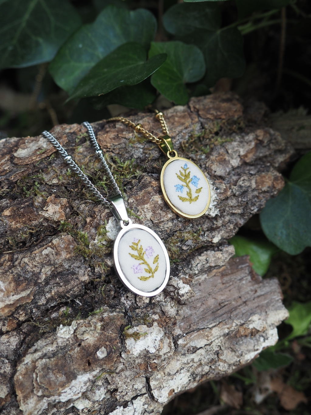 L'image présente deux pendentifs ovales délicats, chacun orné d'un motif floral brodé, posés sur un morceau de bois texturé et recouvert de mousse. Le pendentif au premier plan a un cadre argenté brillant et une base de couleur crème sur laquelle est brodée une petite grappe de fleurs violettes avec des feuilles vertes. Il est attaché à une fine chaîne argentée. Le pendentif à l'arrière-plan a un cadre doré et une base de couleur crème similaire, avec une broderie de fleurs bleues et de feuilles vertes. Il est également attaché à une fine chaîne dorée. Les pendentifs sont positionnés sur un morceau de bois rugueux et écorcé, qui présente des nuances de brun et des textures variées, avec des taches de mousse verte. En arrière-plan, on peut voir des feuilles de lierre vert foncé, créant un contraste naturel et doux avec les bijoux. La lumière semble venir d'en haut, éclairant les pendentifs et le bois, et créant des ombres douces. L'ensemble de la scène évoque une ambiance artisanale et naturelle.