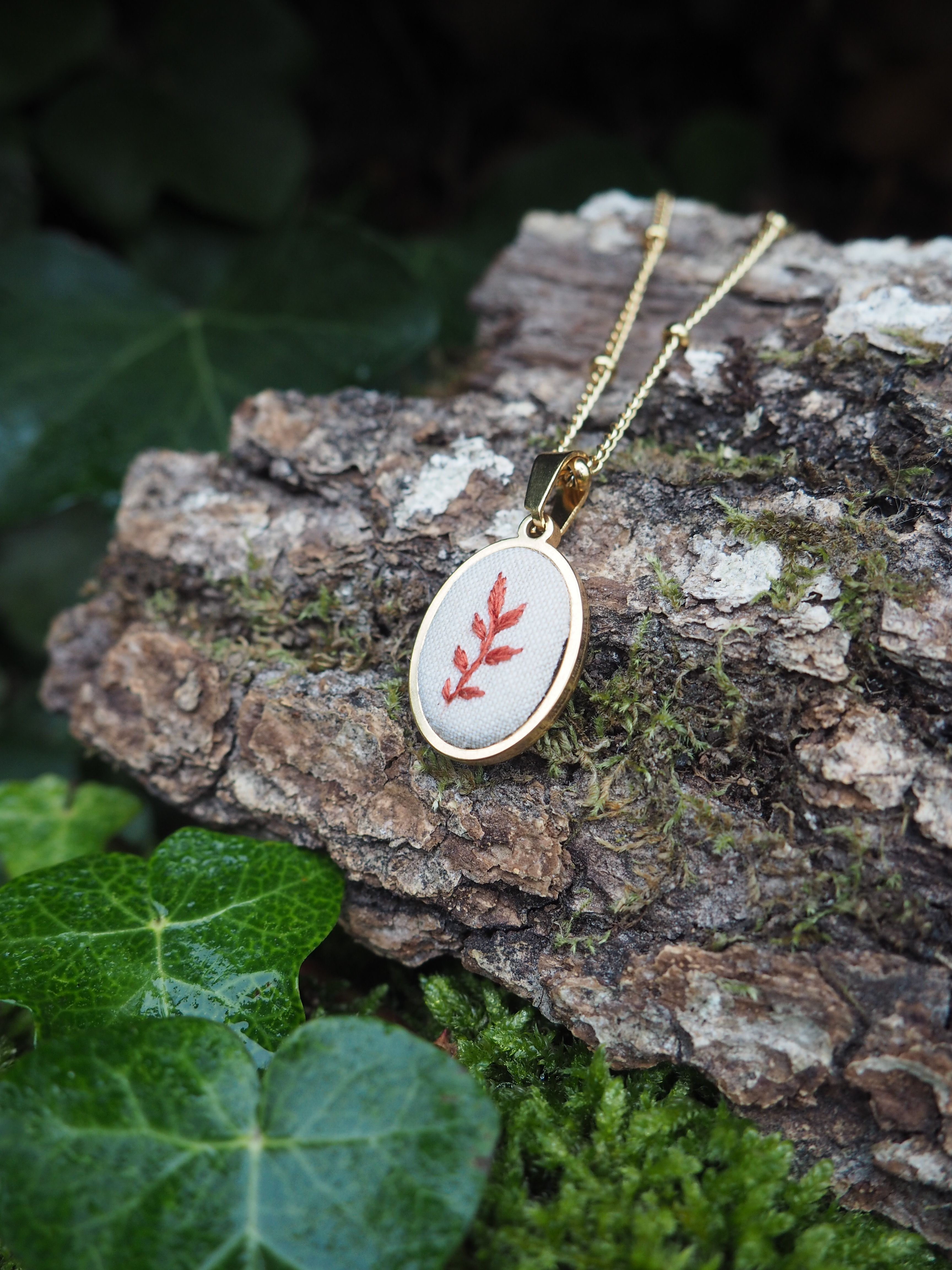 This image features a close-up of a delicate gold-colored necklace with an oval pendant, resting on a piece of tree bark. The pendant has a white fabric insert with a small, reddish-brown embroidered leaf design. The necklace chain is thin and also gold-colored, with small, evenly spaced beads. The tree bark is textured and appears to be covered in patches of green moss, suggesting an outdoor, natural setting. In the foreground and background, there are vibrant green leaves, possibly ivy, some of which appear slightly wet or dewy, adding to the natural and fresh aesthetic of the scene. The lighting is soft, highlighting the details of the necklace and the textures of the natural elements.