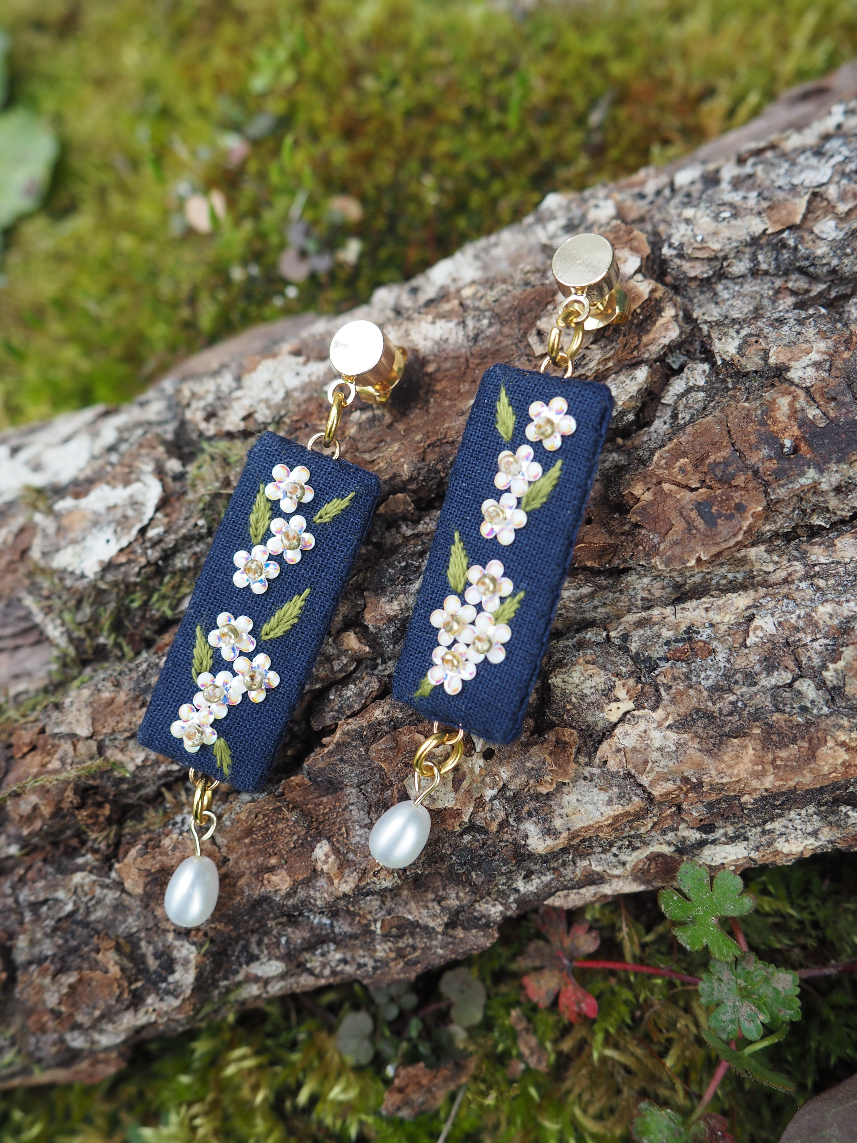 Cette image montre une paire de boucles d'oreilles artisanales élégantes. Elles sont composées d'un rectangle de tissu bleu marine sur lequel sont brodées de délicates fleurs blanches et roses, rappelant des fleurs de cerisier. Chaque boucle d'oreille est ornée d'une perle blanche qui pend au bout. Les boucles d'oreilles sont photographiées sur ce qui semble être un morceau d'écorce d'arbre recouvert de mousse verte, créant un beau contraste naturel avec les bijoux. Le style est très japonisant et délicat, mélangeant des éléments traditionnels (motifs floraux) avec un design contemporain.