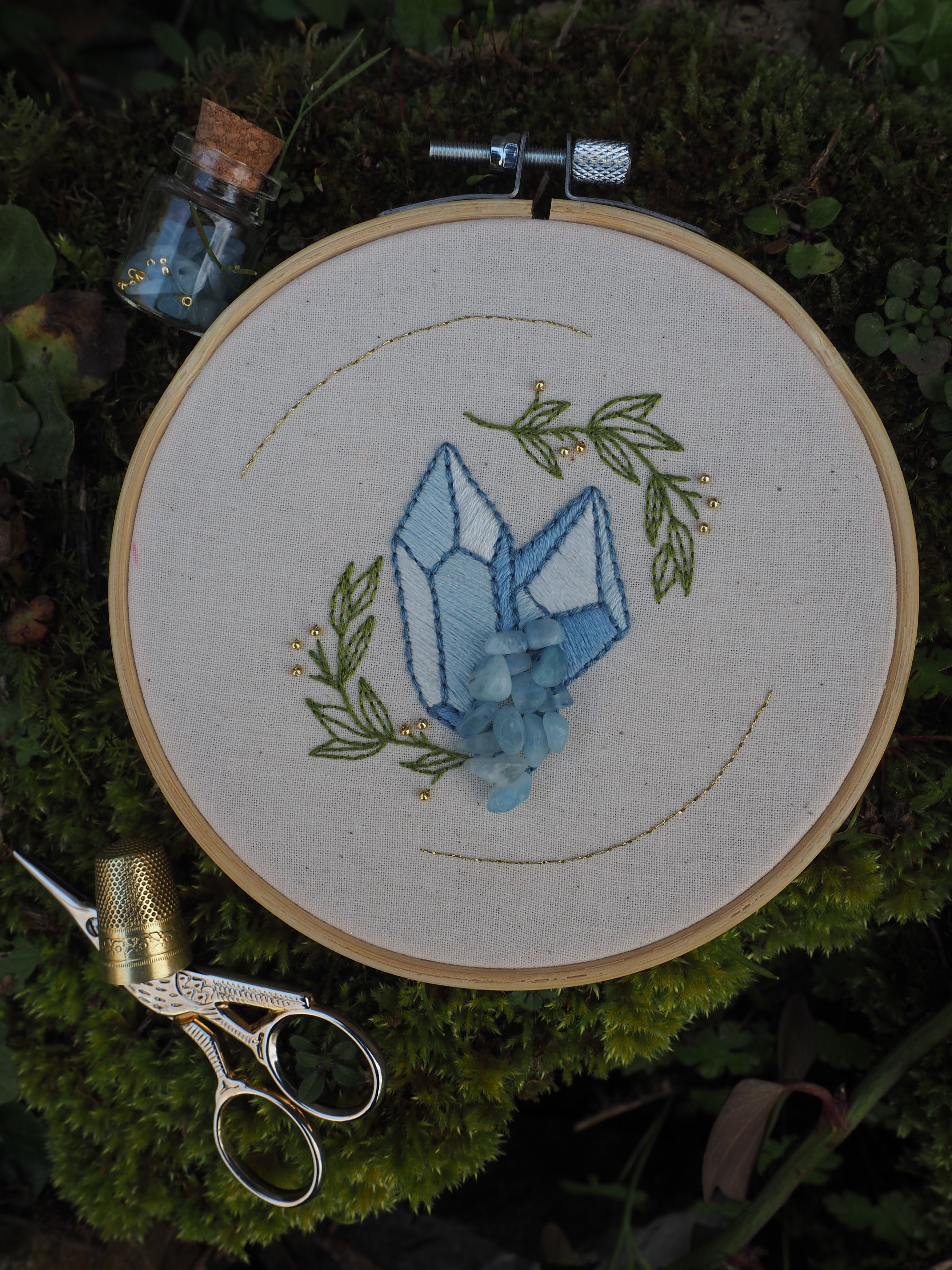 Cette image montre une broderie dans un cercle à broder en bois. Le motif représente un cristal ou une pierre précieuse en bleu clair, entouré de feuilles vertes brodées. La broderie est réalisée sur un tissu blanc ou écru. On peut également voir des ciseaux de broderie dorés et ce qui semble être un petit flacon en verre avec un bouchon en liège dans le cadre de la photo. L'ensemble est photographié sur ce qui semble être de la mousse ou de la végétation verte en arrière-plan. C'est un bel exemple d'artisanat textile avec un design moderne et minimaliste.