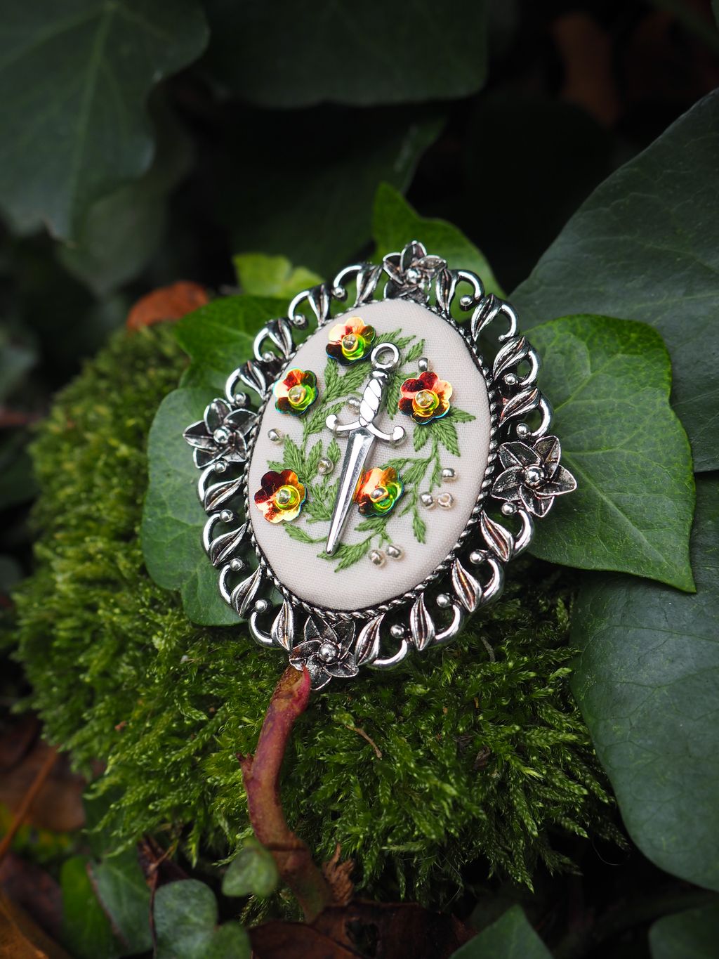 Cette image présente un magnifique bijou artisanal posé sur un lit de mousse et de feuillage naturel. Il s'agit d'une broche ou d'un pendentif de forme circulaire avec un design élaboré : **Caractéristiques du bijou :**
- **Monture** : Un cadre en métal argenté travaillé avec des motifs décoratifs en relief tout autour du pourtour
- **Centre** : Un médaillon en porcelaine ou céramique blanche ornée d'un motif peint à la main représentant des fleurs et du feuillage dans des tons verts
- **Détails colorés** : De petites pierres ou cabochons de couleur ambrée/dorée sont sertis dans la monture métallique
- **Style** : Le bijou évoque l'artisanat traditionnel ou vintage, avec un travail minutieux et raffiné **Mise en scène :**
L'objet est photographié dans un environnement naturel sur de la mousse verte et des feuilles, créant un contraste harmonieux entre l'artifice du bijou et la nature environnante. L'éclairage met en valeur les détails du travail métallique et fait ressortir les couleurs délicates du motif central. Ce type de bijou semble être une pièce d'artisanat de qualité, possiblement inspirée de styles européens traditionnels.