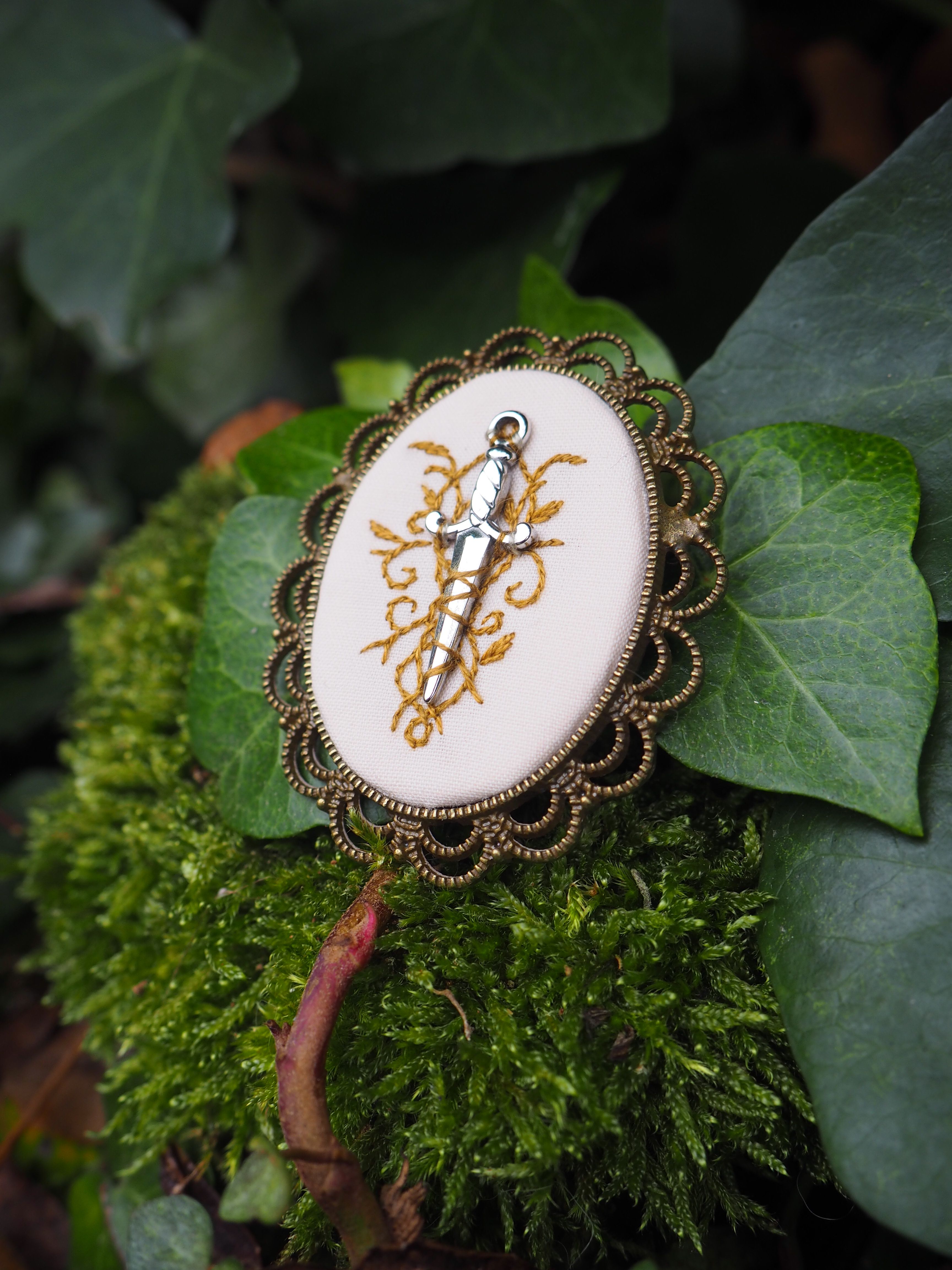 Cette image présente un bijou décoratif posé sur un lit de végétation luxuriante. Il s'agit d'une broche ou d'un pendentif de forme ovale, serti dans un cadre métallique orné de détails ciselés qui lui donnent un aspect vintage ou antique. Le centre du bijou arbore un fond blanc ou crème sur lequel est représenté un motif doré élégant. Ce motif semble être de style baroque ou rococo, avec des courbes gracieuses et des ornements floraux stylisés qui évoquent les arts décoratifs classiques. Le bijou repose sur un tapis de mousse verte d'un vert éclatant, entouré de feuilles de différentes plantes. On peut observer des feuilles aux textures variées - certaines lisses et d'autres plus rugueuses - dans des tons de vert allant du vert tendre au vert plus soutenu. Cette mise en scène naturelle crée un contraste saisissant entre l'objet manufacturé raffiné et l'environnement végétal organique. L'éclairage doux met en valeur les détails du bijou tout en préservant l'atmosphère naturelle et sereine de la composition.