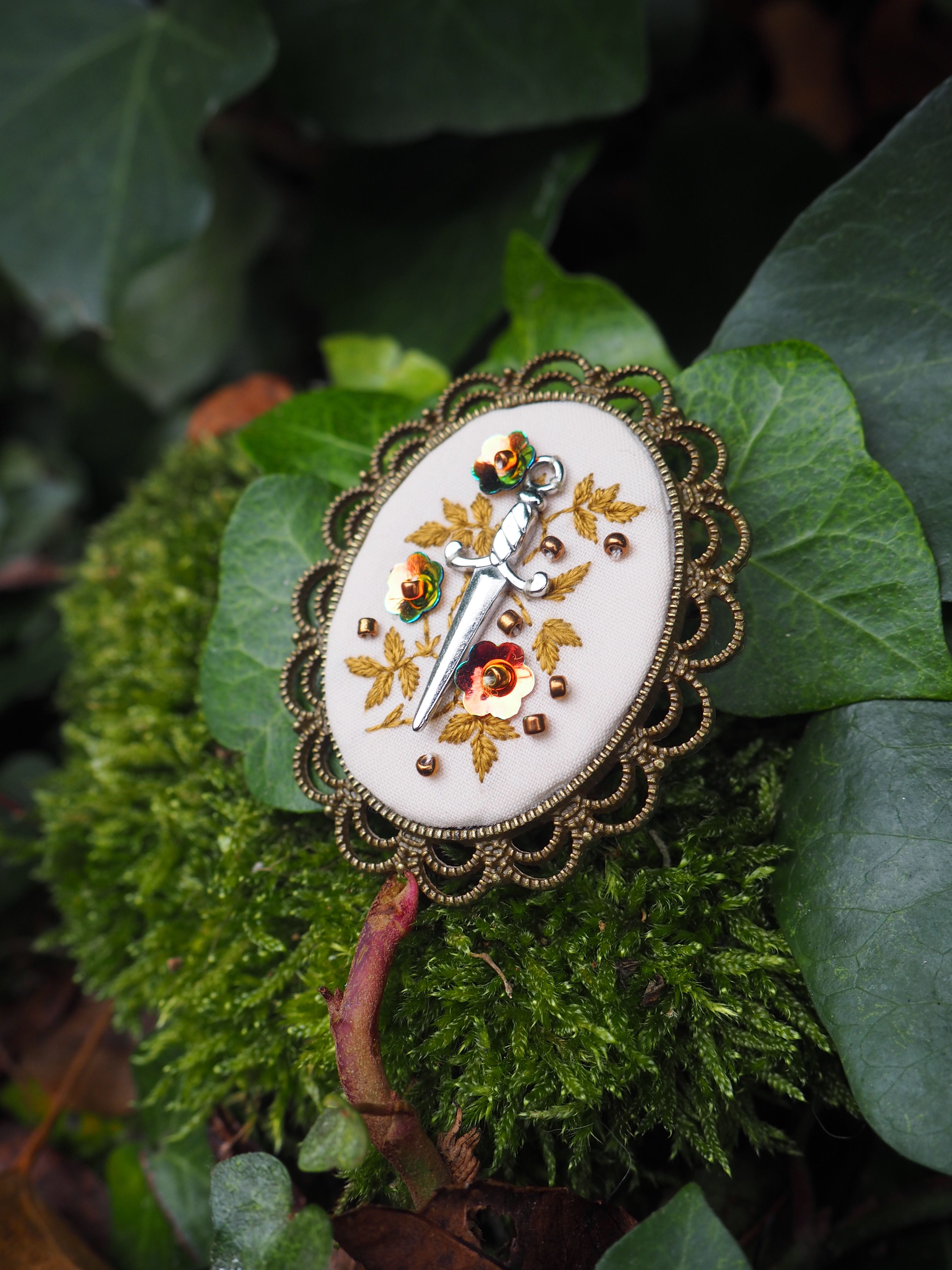 Cette image présente un magnifique pendentif ou broche artisanale posé délicatement sur un lit de mousse verte et de feuillage naturel.  Le bijou se compose d'un médaillon ovale à bordure dorée finement ciselée avec des motifs décoratifs en relief. Au centre, on découvre une miniature peinte représentant un oiseau aux couleurs vives - probablement un rouge-gorge ou un oiseau similaire - perché sur une branche ornée de feuilles dorées et de petites baies rouges. Le style de cette peinture évoque l'art décoratif traditionnel européen ou les techniques de miniature sur porcelaine. L'ensemble repose sur un écrin naturel composé de mousse d'un vert intense et de grandes feuilles, créant un contraste saisissant entre l'objet artisanal raffiné et son environnement végétal sauvage. Cette mise en scène photographique met en valeur à la fois la délicatesse du travail d'orfèvrerie et la beauté de la nature qui l'entoure. L'éclairage doux fait ressortir les détails dorés du bijou tout en préservant les nuances subtiles du feuillage environnant.