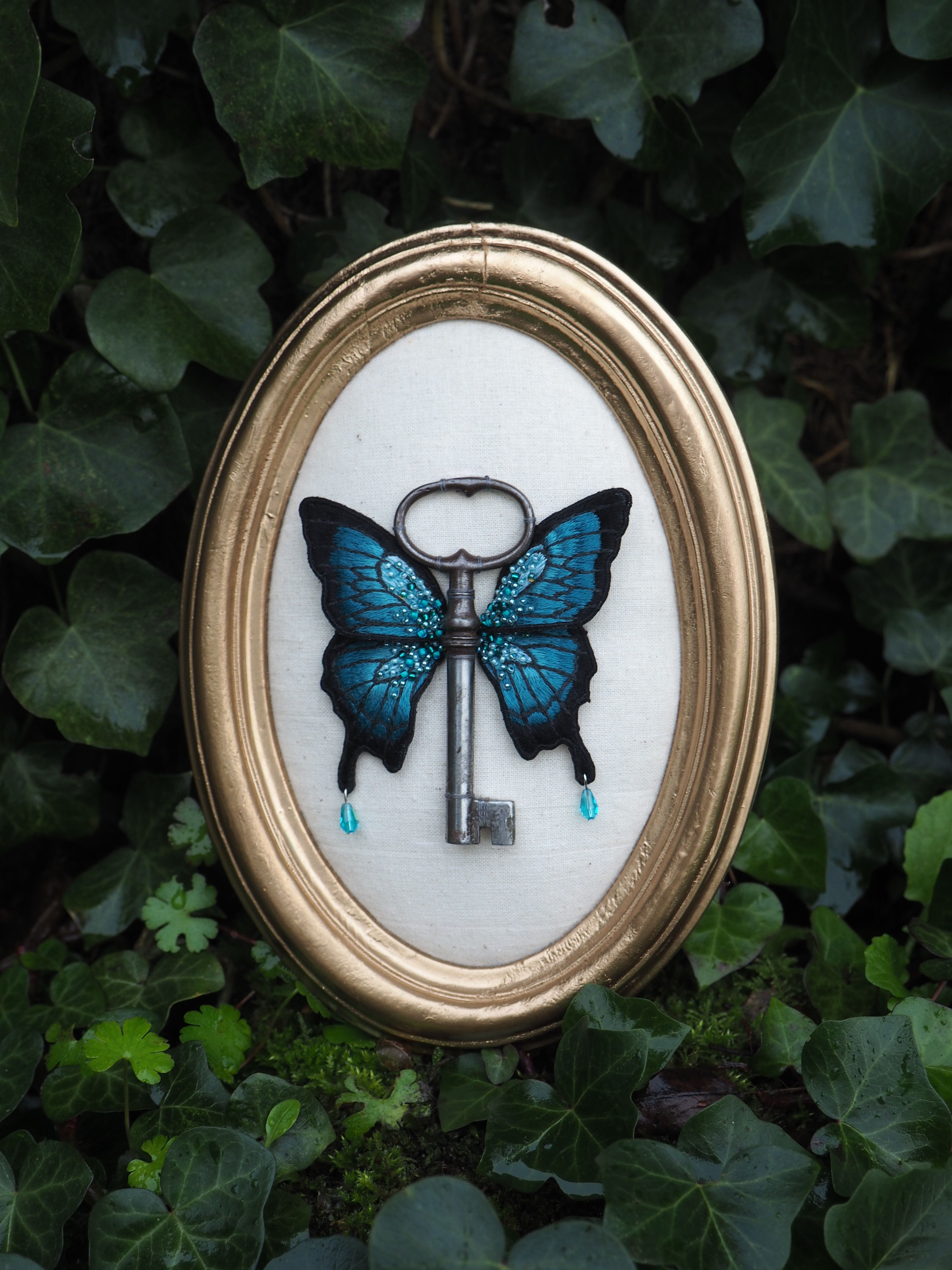 Cette image montre une œuvre d'art encadrée très créative et symbolique. Elle présente une clé antique dont les ailes sont formées par les ailes d'un papillon bleu brillant. La pièce est présentée dans un cadre rond doré classique et est placée contre un fond de feuilles de lierre vert. Les ailes du papillon sont d'un bleu vibrant avec des motifs délicats, et deux petites gouttes bleues semblent tomber de chaque côté de la clé. L'ensemble crée une composition poétique et mystérieuse qui évoque la liberté, la transformation et peut-être même un passage secret ou magique. Le contraste entre le métal de la clé et les ailes délicates du papillon crée une juxtaposition intéressante entre le dur et le délicat, le pratique et le fantaisiste.
