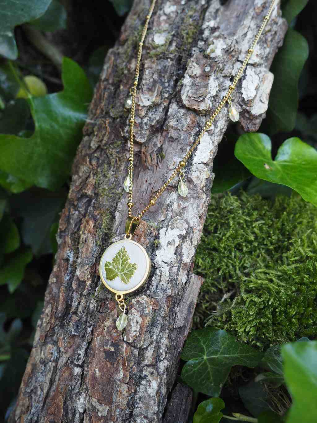 L'image présente un collier délicat et artisanal, suspendu sur un morceau de bois brut, probablement une branche ou un tronc d'arbre. Le collier est composé d'une fine chaîne dorée, ornée de petites perles translucides de couleur vert pâle, qui pendent à intervalles réguliers le long de la chaîne. Le pendentif central est rond, avec un cadre doré qui entoure une surface blanche ou crème. Au centre de ce pendentif, une petite feuille verte, vraisemblablement une feuille de lierre ou une feuille similaire, est incrustée ou représentée, créant un contraste doux et naturel avec le fond clair. Sous le pendentif principal, une petite grappe de perles translucides similaires à celles de la chaîne est suspendue, ajoutant un mouvement et une touche d'éclat. Le fond est composé d'éléments naturels. Le morceau de bois sur lequel repose le collier a une texture rugueuse et écorcée, avec des nuances de brun et de gris. Autour du bois, on peut voir du feuillage vert foncé, probablement du lierre, qui s'entrelace et ajoute de la profondeur à la composition. Sur la droite, une zone de mousse verte et dense est visible, renforçant l'ambiance naturelle et forestière de la scène. L'éclairage semble naturel, mettant en valeur les détails du collier et les textures des éléments environnants. L'ensemble de l'image dégage une atmosphère douce, organique et artisanale, suggérant un bijou inspiré par la nature.