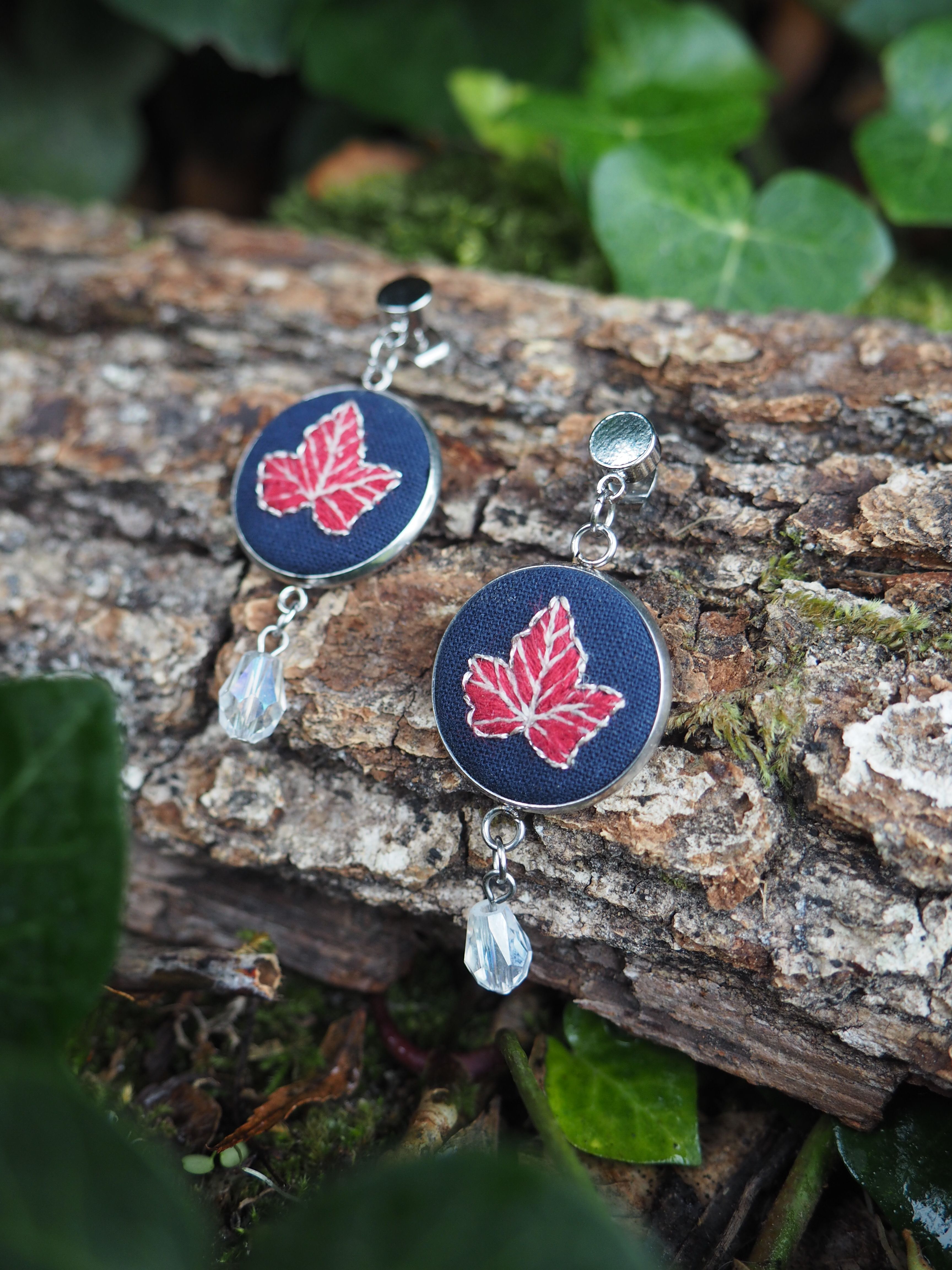 Cette image présente une paire de boucles d'oreilles artisanales disposées sur un tronc d'arbre dans un environnement naturel.  **Caractéristiques des bijoux :**
- Forme circulaire avec un fond noir mat
- Motif central : feuille d'érable stylisée en rouge/rose
- Finition argentée pour les crochets et bordures
- Petites perles transparentes ou cristallines suspendues en pendentif
- Style artisanal avec une esthétique moderne **Mise en scène :**
- Posées sur l'écorce rugueuse d'un tronc d'arbre
- Environnement forestier avec de la mousse verte visible
- Feuillage naturel en arrière-plan créant un bokeh doux
- Éclairage naturel mettant en valeur les détails L'ensemble crée un contraste harmonieux entre l'artisanat humain et la nature environnante, soulignant le thème de la feuille d'érable qui fait écho à l'environnement forestier. La photographie met bien en valeur la qualité artisanale de ces bijoux aux couleurs automnales.