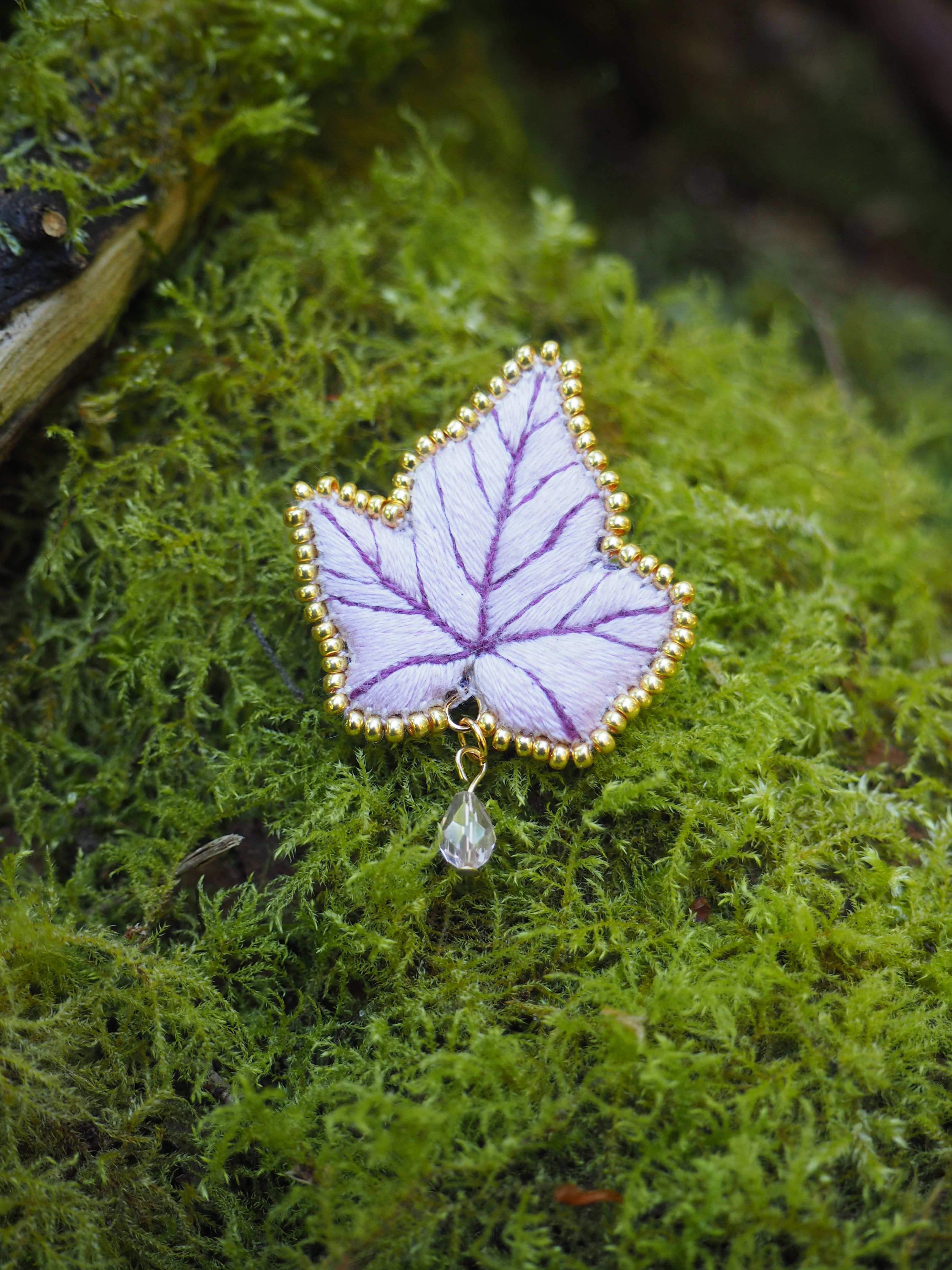 Cette image montre une délicate broche ou un pendentif en forme de feuille posé sur un lit de mousse verte. Le bijou est fait dans des tons de violet et blanc, avec des veines violettes qui rappellent celles d'une véritable feuille. Les bords sont ornés de petites perles dorées qui forment une bordure décorative. Un petit cristal ou une perle est suspendu en bas de la feuille. Le contraste entre le bijou délicat et la mousse naturelle verte crée une composition très poétique et féerique.