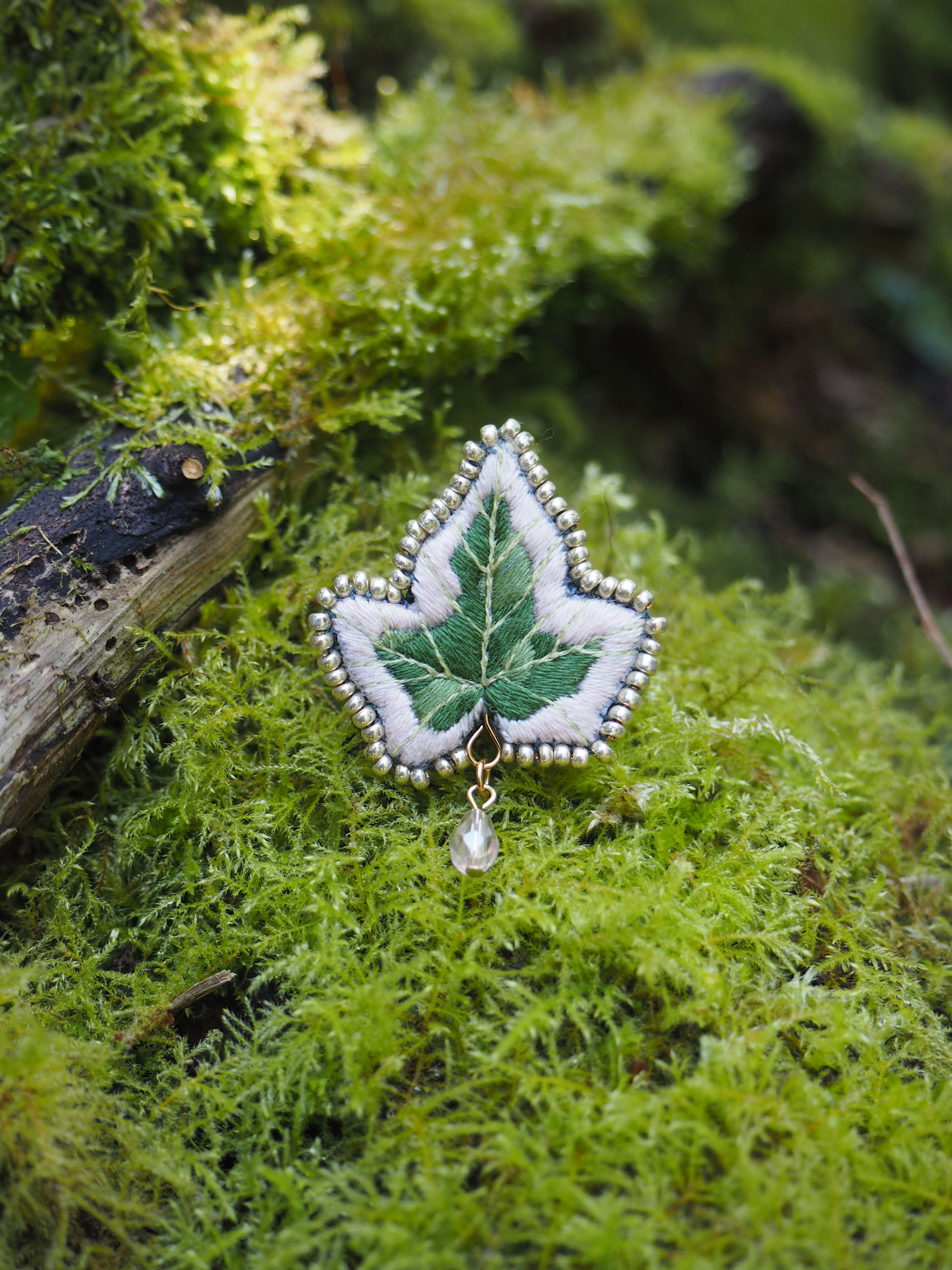 Cette image montre une belle broche ou un pendentif en forme de feuille d'érable sur un fond de mousse verte luxuriante. Le bijou semble être fait d'émail vert et blanc, avec des perles ou des petites pierres blanches bordant le contour de la feuille. Le design est délicat et détaillé, capturant parfaitement la forme caractéristique de la feuille d'érable. Une petite perle ou pierre est suspendue au bas du bijou. La composition est très naturelle, avec le bijou posé sur un lit de mousse verte et fraîche, créant un beau contraste entre l'objet artisanal et l'environnement naturel.