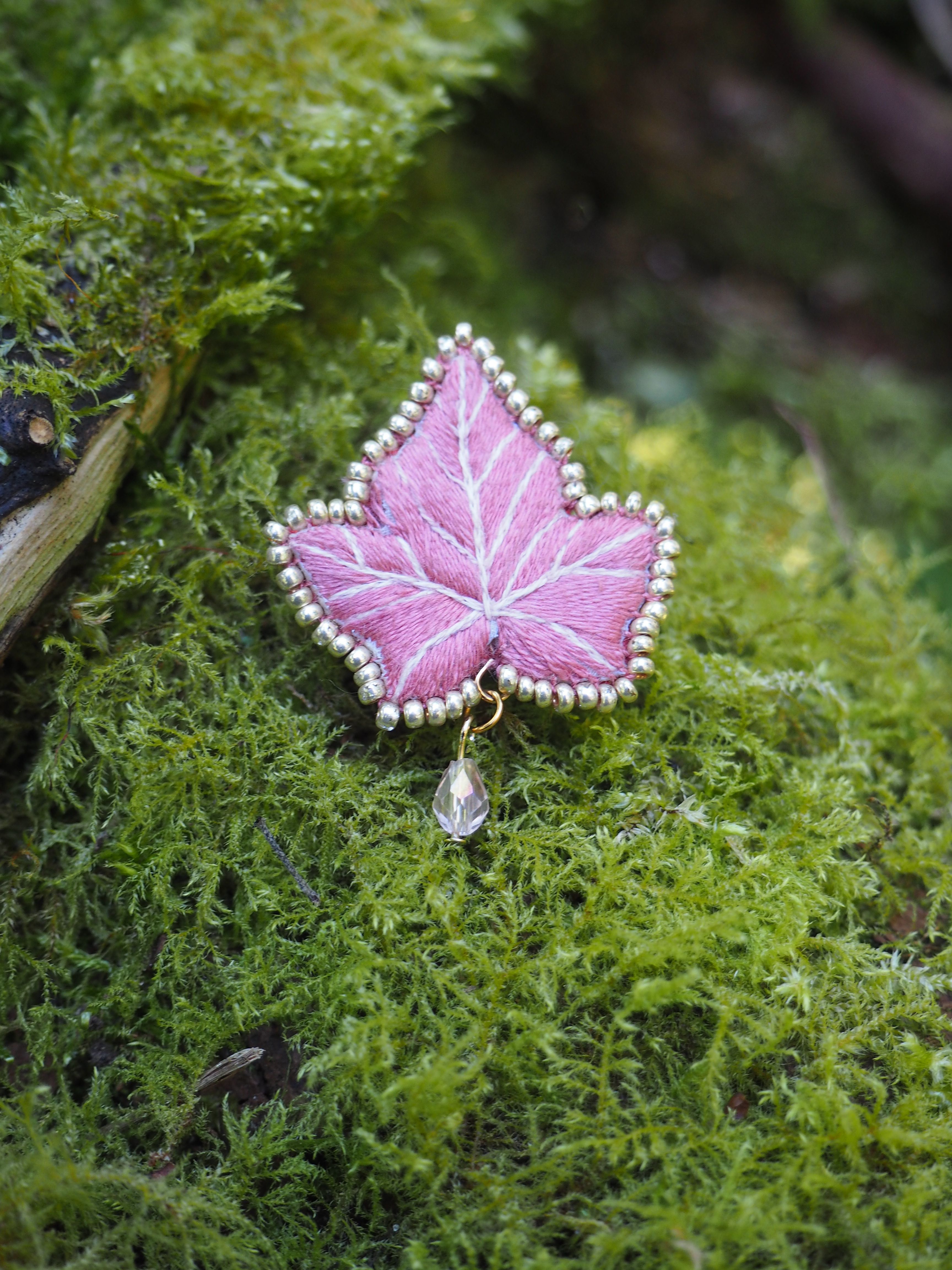 Cette image montre un délicat bijou en forme de feuille d'érable posé sur un lit de mousse verte. Le bijou est d'une belle couleur rose avec des détails de nervures blanches, et son contour est orné de petites perles argentées qui lui donnent un aspect élégant. Un petit cristal ou une perle est suspendu au bas de la feuille. Le contraste entre le rose doux du bijou et le vert vif de la mousse crée une composition très esthétique et naturelle. C'est une photographie qui capture parfaitement la rencontre entre un élément artificiel raffiné et la texture naturelle de la mousse.
