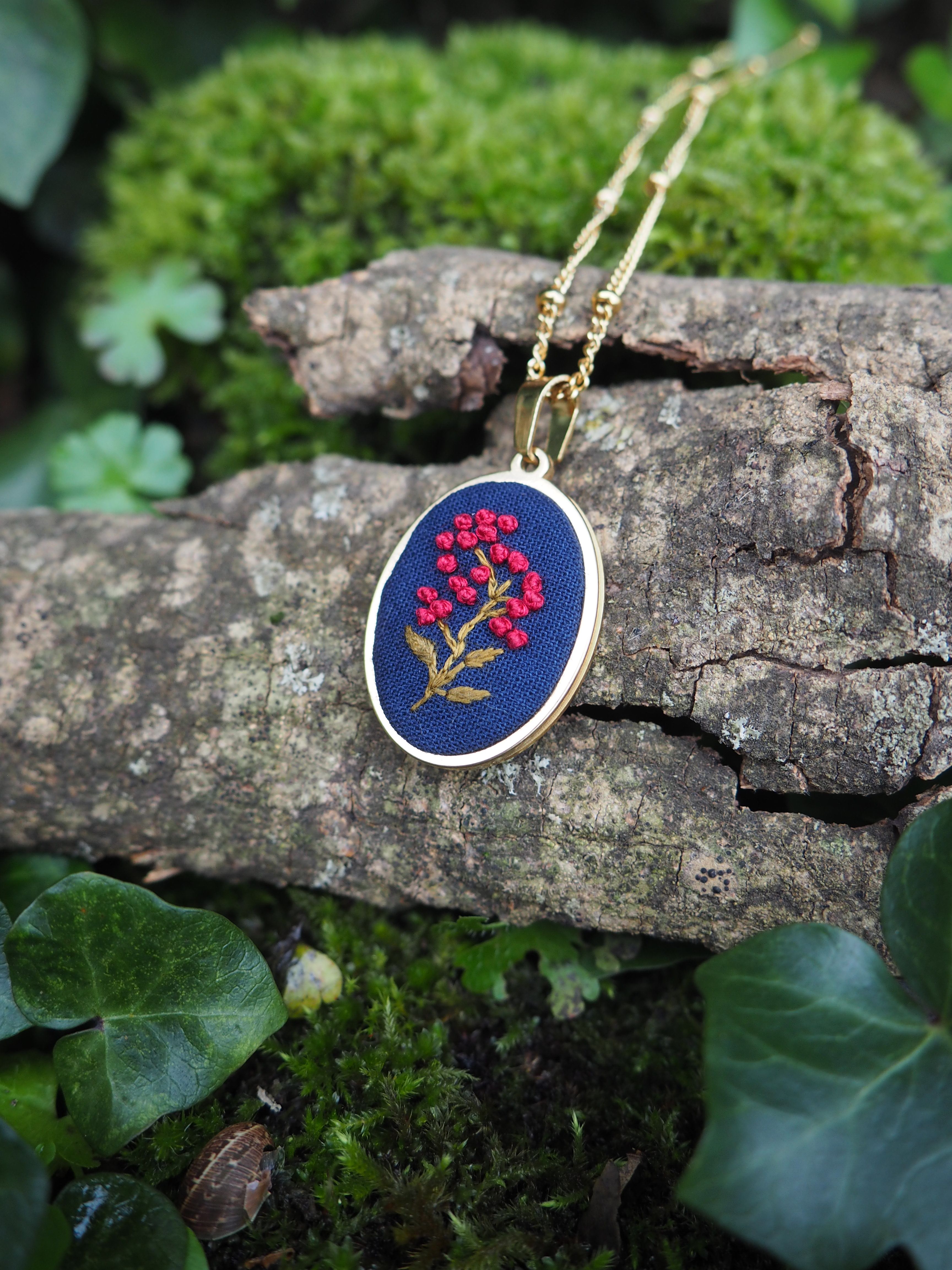 L'image montre un pendentif délicat posé sur un morceau de bois ou d'écorce dans un environnement naturel. Le pendentif est rond, avec un fond bleu foncé sur lequel est brodé un motif floral rouge et doré. La broderie représente une petite branche ou une fleur avec des pétales rouges et une tige dorée. Le pendentif est suspendu à une fine chaîne dorée, probablement en or. Il repose sur ce qui semble être un morceau de bois ou d'écorce texturé et gris, entouré de mousse verte et de feuilles. Cette mise en scène naturelle met en valeur la délicatesse et la beauté artisanale du bijou. L'arrière-plan flou montre des touches de vert, suggérant un cadre forestier ou un jardin. L'ensemble de la composition crée une ambiance naturelle et romantique, mettant en valeur le contraste entre le bijou raffiné et son environnement rustique.