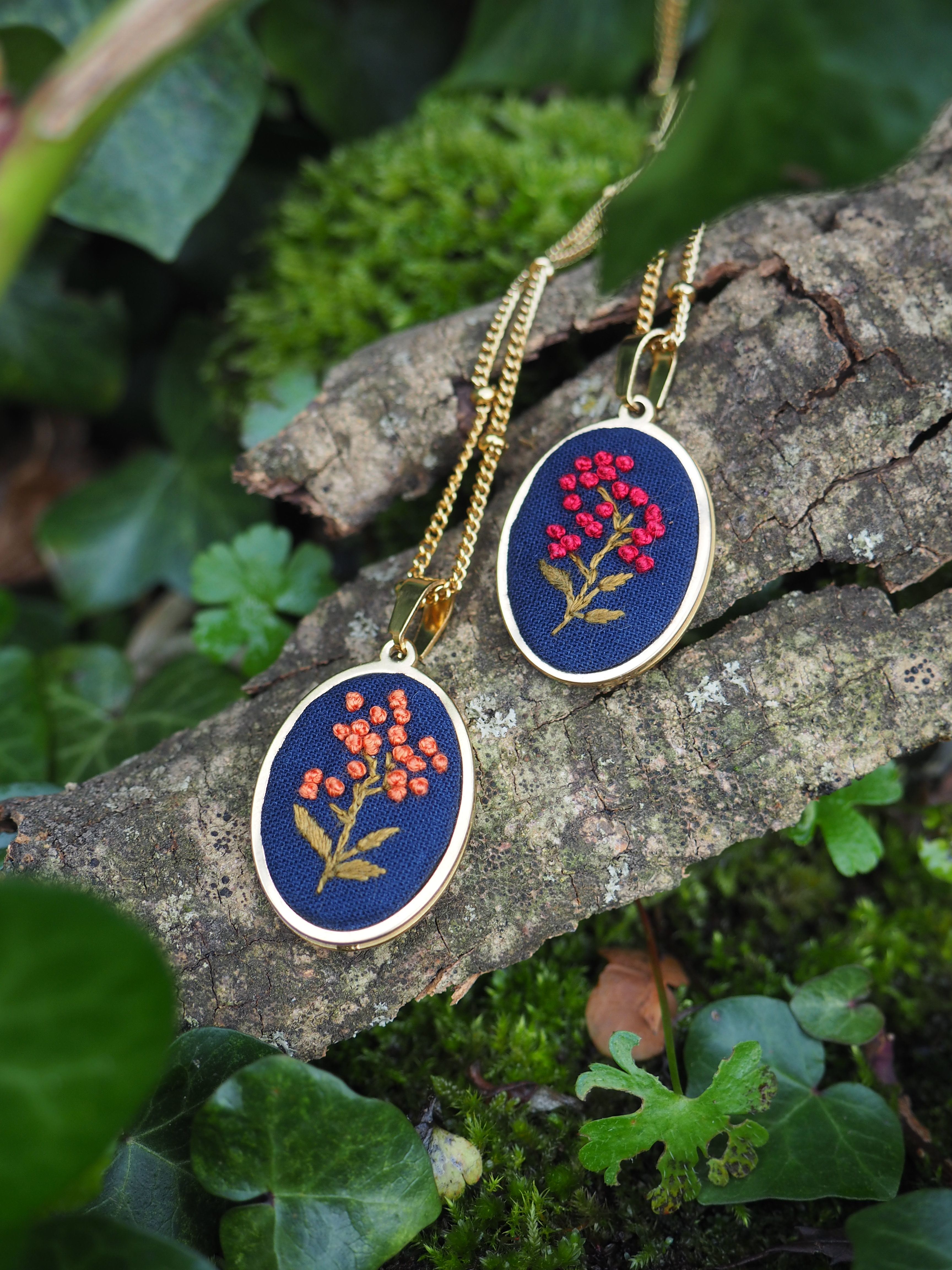 L'image montre deux beaux pendentifs brodés posés sur un morceau de bois ou d'écorce, entourés de feuilles vertes. Chaque pendentif est un médaillon rond avec un fond bleu foncé, sur lequel est brodé un motif floral délicat en rouge et or. Les fleurs ressemblent à de petites grappes de baies ou de fleurs, avec des tiges et des feuilles dorées. Les pendentifs sont suspendus à des chaînes dorées, ce qui suggère qu'il s'agit de colliers. Le cadre naturel, avec le bois texturé et la végétation verte en arrière-plan, met en valeur la beauté artisanale de ces bijoux. Ces pendentifs semblent être des pièces de bijouterie faites main, probablement créées en utilisant des techniques de broderie traditionnelles. Leur design est à la fois simple et élégant, combinant des couleurs vives avec un fond sombre pour un effet saisissant. Ce type de bijou pourrait être apprécié par ceux qui aiment les accessoires uniques et artisanaux avec une touche naturelle ou florale.