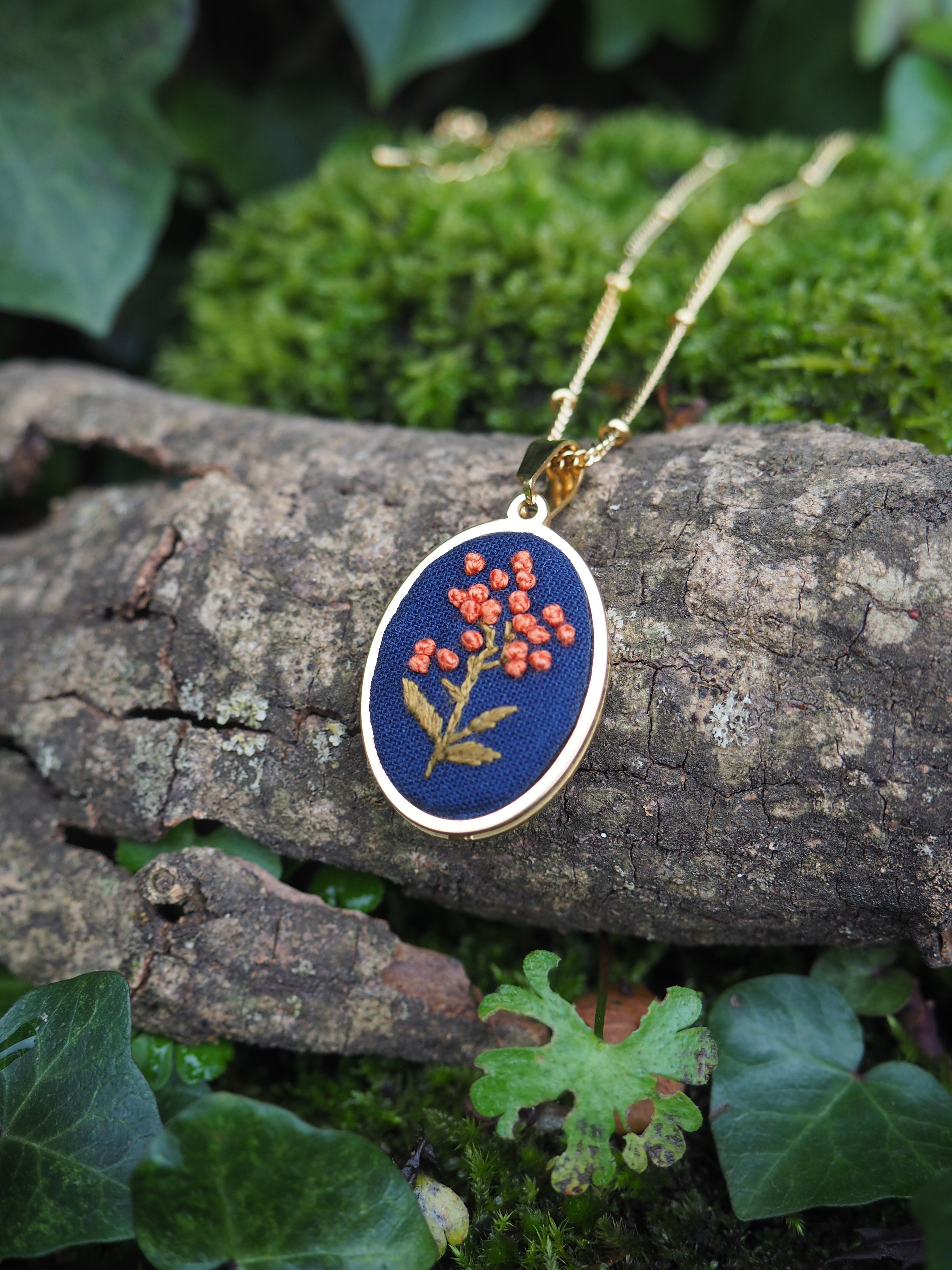 L'image montre un joli pendentif de broderie posé sur un morceau de bois ou d'écorce. Le pendentif est rond, avec un fond bleu foncé sur lequel est brodé un motif floral délicat en fils oranges et rouges. La fleur brodée ressemble à une petite branche avec des baies ou des fleurs. Le pendentif est attaché à une fine chaîne dorée, suggérant qu'il s'agit d'un collier. L'ensemble est placé dans un cadre naturel, avec de la mousse verte et des feuilles de lierre visibles autour du morceau de bois. Cette composition crée un contraste intéressant entre l'objet artisanal fait main et l'environnement naturel, mettant en valeur la beauté du travail de broderie tout en le situant dans un contexte organique et verdoyant. L'image a une qualité esthétique qui évoque la délicatesse et la connexion avec la nature.