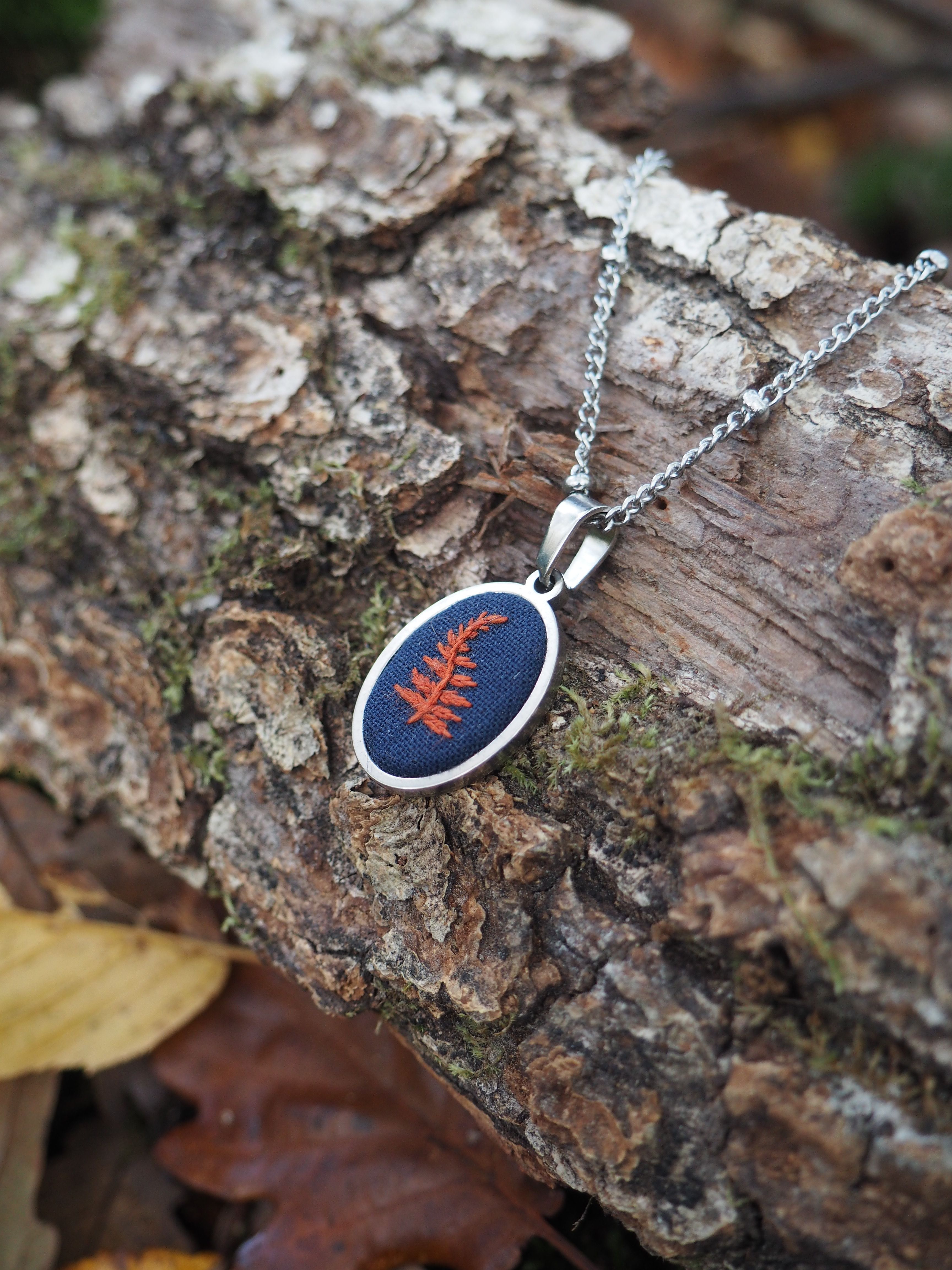 Cette image présente un pendentif élégant posé sur un morceau d'écorce naturelle dans un environnement forestier. Le bijou se compose d'une chaîne argentée délicate reliée à un pendentif de forme ovale avec un cadre métallique argenté. Le pendentif contient une résine ou un matériau transparent de couleur bleue profonde dans lequel est enchâssée une feuille naturelle de couleur rouge-orangé, probablement une fougère ou une petite feuille d'automne. Cette inclusion végétale crée un contraste saisissant avec le fond bleu. L'ensemble repose sur une surface d'écorce rugueuse et texturée, avec des tons bruns et gris naturels. On peut apercevoir quelques mousses verdâtres sur l'écorce, ainsi qu'une feuille jaunâtre en arrière-plan, ce qui renforce l'ambiance naturelle et organique de la composition. L'éclairage met bien en valeur les détails du bijou et crée une atmosphère chaleureuse qui souligne le thème nature de cette pièce artisanale. Ce type de pendentif semble être un bijou artisanal qui capture et préserve un élément naturel dans une résine colorée.