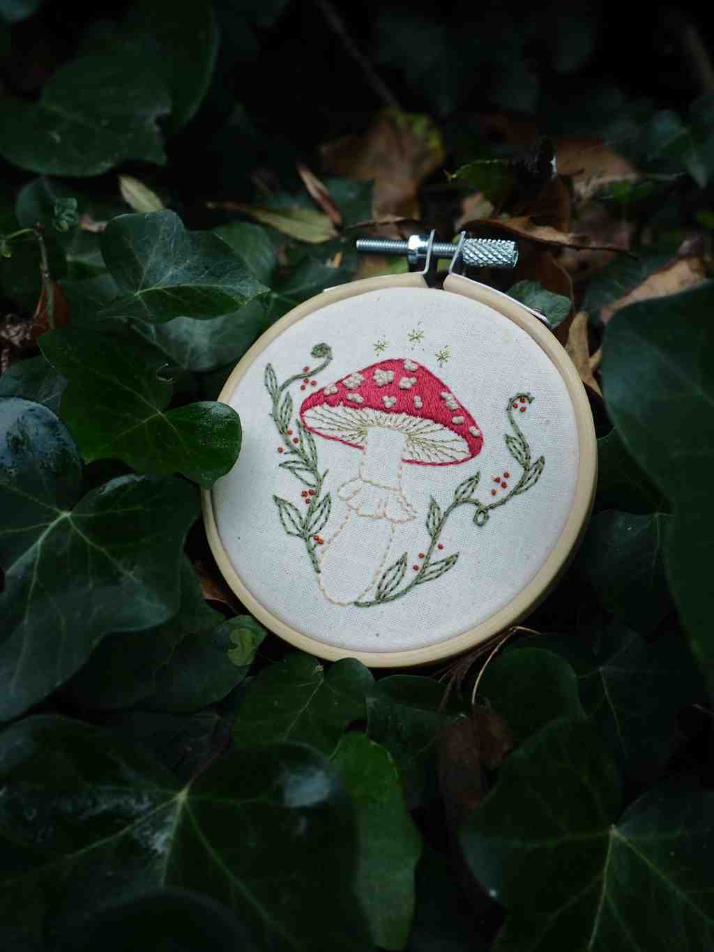 Cette image présente une charmante broderie artisanale disposée dans un environnement naturel. Voici ce que je peux observer : **L'objet principal :**
- Un tambour à broder circulaire en bois contenant une broderie sur tissu blanc
- Le motif brodé représente un champignon rouge à pois blancs (probablement une amanite tue-mouches) avec sa tige blanche
- Le champignon est entouré de délicates branches et feuillages brodés en vert
- Le travail de broderie semble fait à la main avec différents points et couleurs **Le cadre naturel :**
- Le tambour est posé sur un lit de feuilles vertes, créant un contraste harmonieux
- L'environnement semble être de la végétation dense, possiblement du lierre ou des feuilles similaires
- L'éclairage naturel met en valeur les détails de la broderie
- La composition crée un lien poétique entre l'art textile et la nature Cette mise en scène évoque l'univers des contes de fées et souligne le lien entre l'artisanat traditionnel et les motifs inspirés de la nature. La qualité artistique de la broderie et sa présentation dans ce cadre végétal créent une atmosphère à la fois nostalgique et enchantée.