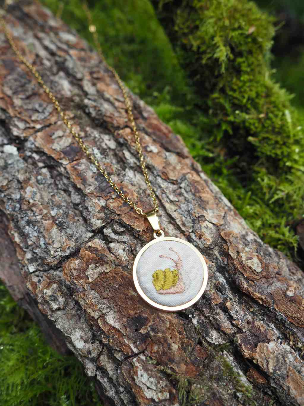L'image présente un collier délicat posé sur un morceau d'écorce d'arbre texturée, avec un arrière-plan flou de mousse verte. Le collier est composé d'une fine chaîne dorée et d'un pendentif circulaire. Le pendentif a un cadre doré brillant et contient une petite broderie ou illustration d'un escargot. L'escargot est représenté avec une coquille jaune-vert et un corps rose pâle, sur un fond clair, probablement blanc ou beige. L'écorce d'arbre sur laquelle repose le collier est rugueuse et présente des motifs et des textures naturels, allant du gris au brun foncé. L'arrière-plan est dominé par une mousse verte luxuriante et floue, ce qui crée un contraste doux et naturel avec les couleurs du collier et de l'écorce. La lumière semble venir d'en haut, illuminant le pendentif et la chaîne, et créant de légères ombres sur l'écorce. L'ensemble de l'image dégage une atmosphère naturelle et artisanale, mettant en valeur le bijou.