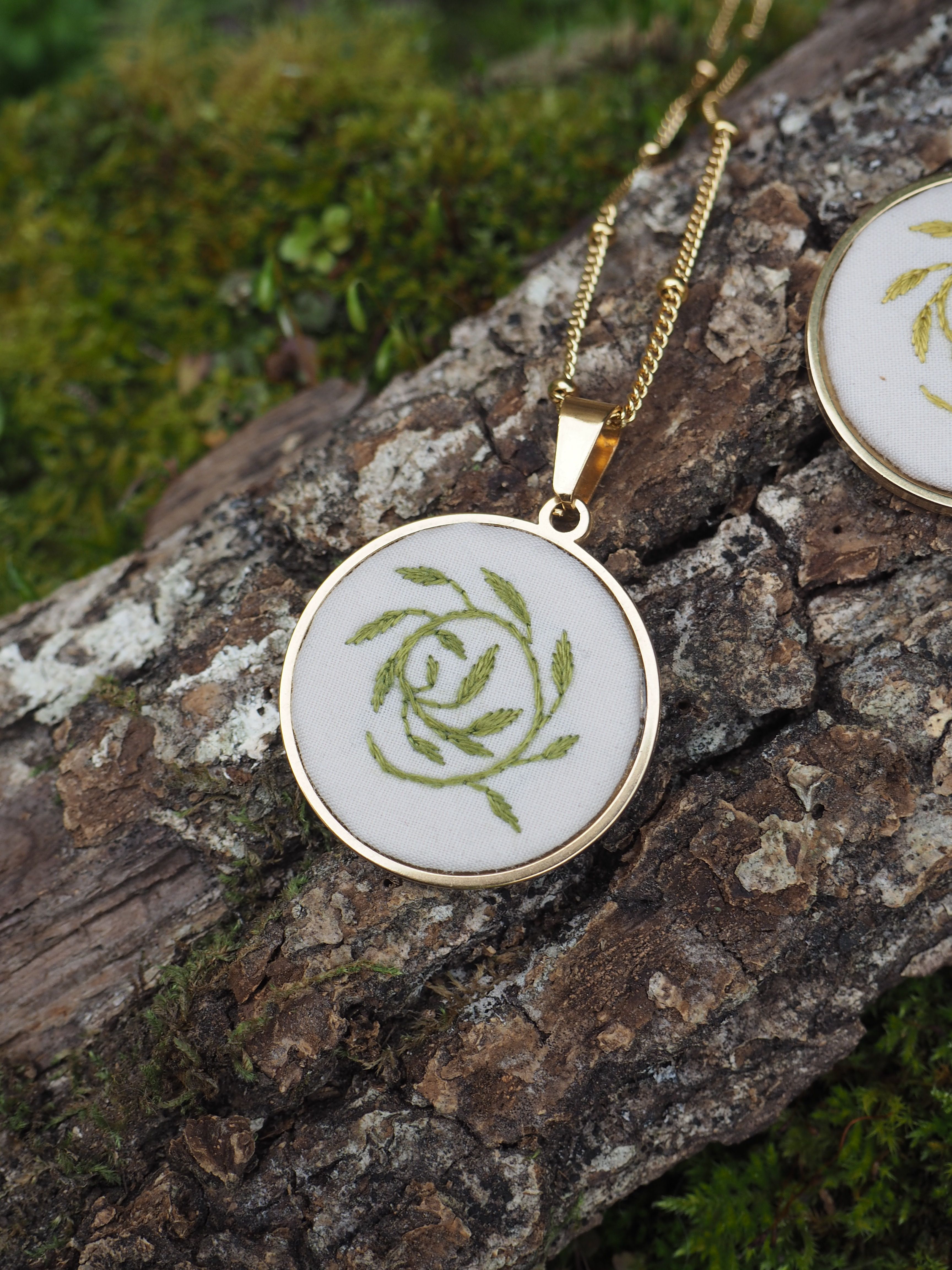 Cette image montre un pendentif élégant posé sur un morceau d'écorce d'arbre recouvert de mousse. Le pendentif est rond, avec un cadre doré, et contient un motif floral vert stylisé ressemblant à une rose sur un fond blanc. Il est attaché à une chaîne dorée fine de style maille. La composition de la photo met en valeur le contraste entre le bijou raffiné et la texture naturelle rugueuse de l'écorce, avec la mousse verte qui fait écho au motif vert du pendentif.