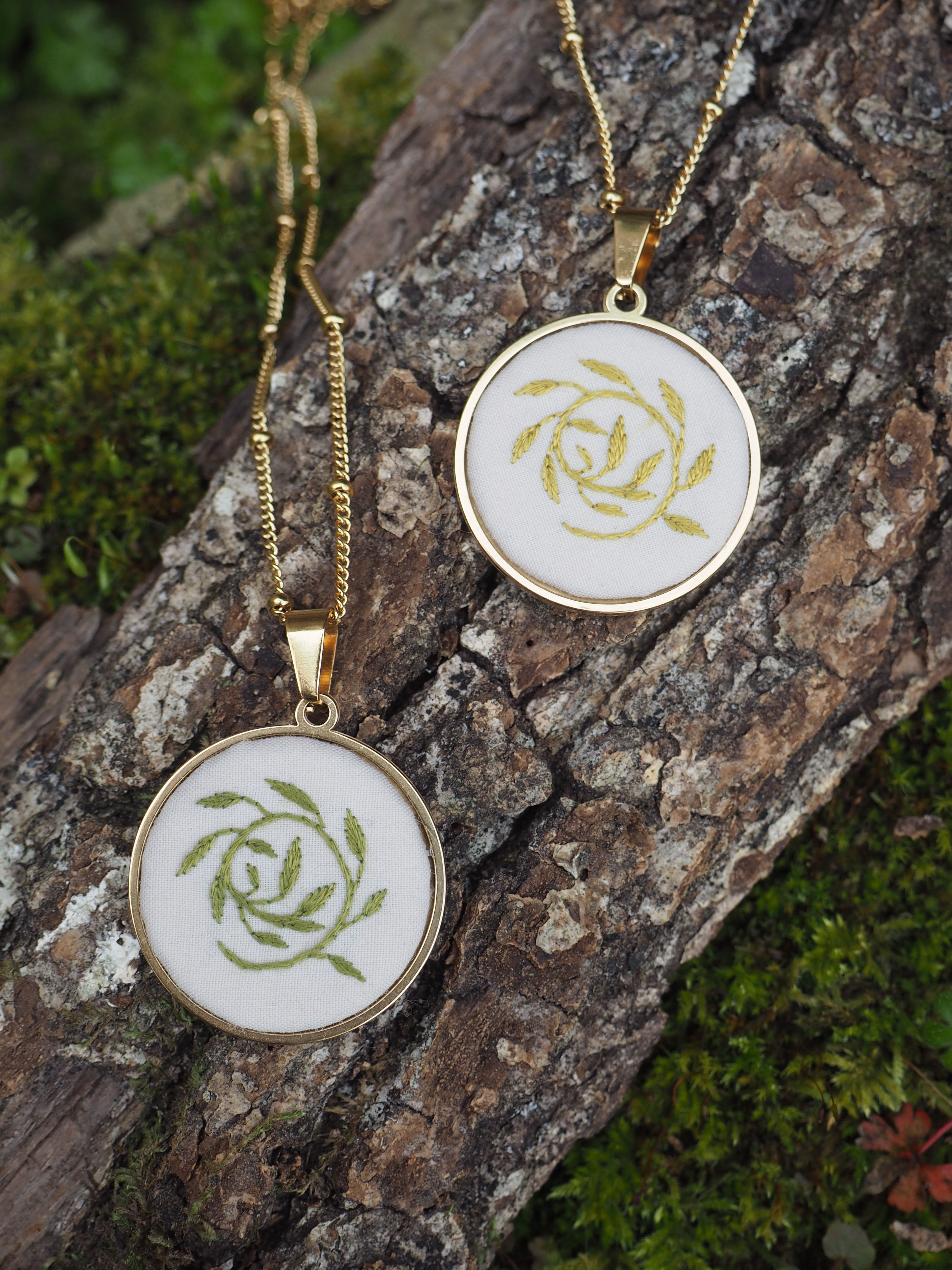 L'image montre deux pendentifs identiques posés sur un morceau de bois ou d'écorce couvert de mousse. Les pendentifs sont constitués de médaillons ovales ou circulaires avec un motif de rose stylisée brodé ou imprimé en vert et or sur un fond blanc. Ils sont montés sur des chaînes dorées. Le cadre des médaillons semble également être en métal doré. La composition est très naturelle et artistique, avec le contraste entre les bijoux délicats et la texture rugueuse du bois et la mousse verte en arrière-plan.