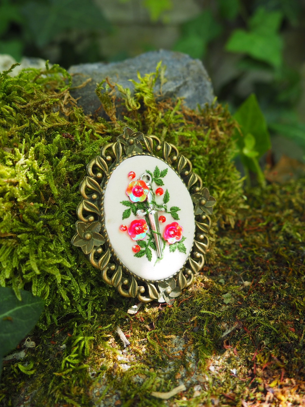 Cette image présente un médaillon ovale orné d'un motif floral et d'une paire de ciseaux, posé sur une surface recouverte de mousse verte. **Description détaillée :** *   **Le médaillon :** Il s'agit d'un médaillon de forme ovale, encadré d'un bord métallique ouvragé de couleur bronze ou laiton vieilli. Le cadre présente des motifs floraux stylisés et des perles en relief, lui donnant un aspect vintage ou antique.
*   **Le motif central :** Au centre du médaillon, sur un fond blanc cassé ou crème, se trouve une broderie ou une peinture miniature. Le motif principal est une paire de ciseaux de couture, représentée de manière réaliste, avec des poignées dorées et des lames argentées. Autour des ciseaux, il y a un arrangement de fleurs et de feuilles. Les fleurs sont principalement des roses ou des bourgeons de roses, de couleur rouge vif et orange, avec des feuilles vertes. L'ensemble du motif est délicat et détaillé.
*   **L'arrière-plan :** Le médaillon est posé sur une surface naturelle, principalement recouverte de mousse verte luxuriante. La mousse est épaisse et texturée, avec des nuances de vert clair et foncé. On peut distinguer quelques petites brindilles ou débris végétaux incrustés dans la mousse.
*   **L'environnement :** À l'arrière-plan, on aperçoit des éléments flous qui suggèrent un environnement extérieur, probablement un jardin ou une forêt. Il y a des zones d'ombre et de lumière, indiquant une source de lumière naturelle, peut-être le soleil filtrant à travers les feuilles. Une feuille d'une plante grimpante (peut-être du lierre) est visible sur le côté droit.
*   **Lumière et ambiance :** La lumière semble naturelle et douce, créant des reflets subtils sur le métal du médaillon et mettant en valeur les couleurs vives du motif floral. L'ambiance générale est calme, naturelle et un peu nostalgique, évoquant l'artisanat et la nature. En résumé, l'image met en scène un beau médaillon artisanal avec un motif de ciseaux et de fleurs, posé délicatement sur un lit de mousse, dans un cadre naturel et lumineux.