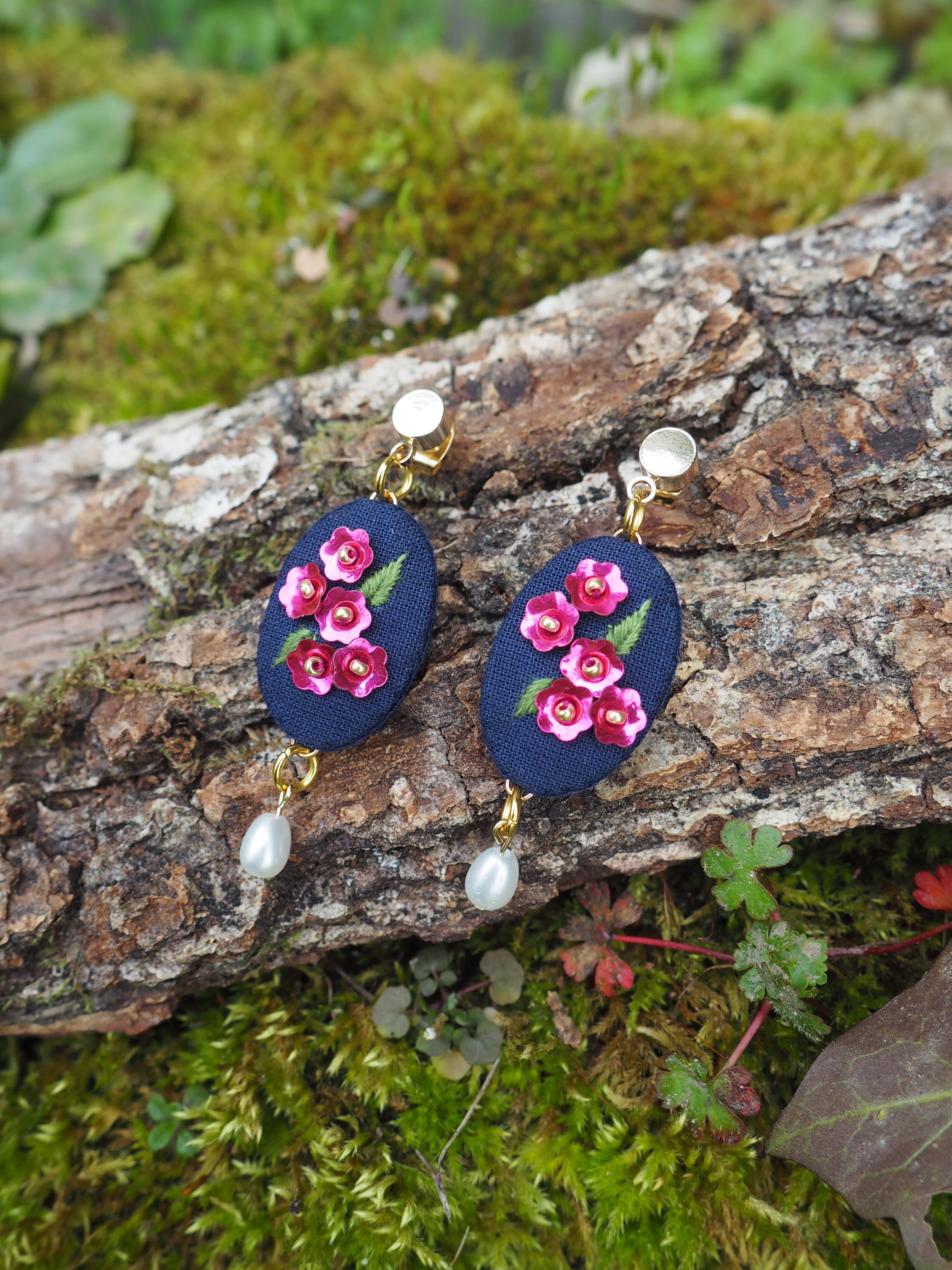 Cette image montre une paire de boucles d'oreilles artisanales posées sur un morceau de bois couvert de mousse. Les boucles d'oreilles sont composées d'un support ovale en tissu bleu marine, décorées de petites fleurs roses brodées. Elles sont agrémentées de perles blanches et de détails dorés. Le contraste entre les bijoux délicats et le fond naturel (bois et mousse verte) crée une belle composition photographique. Ces boucles d'oreilles semblent être des pièces faites main qui combinent élégamment un style romantique avec des éléments naturels comme les perles.