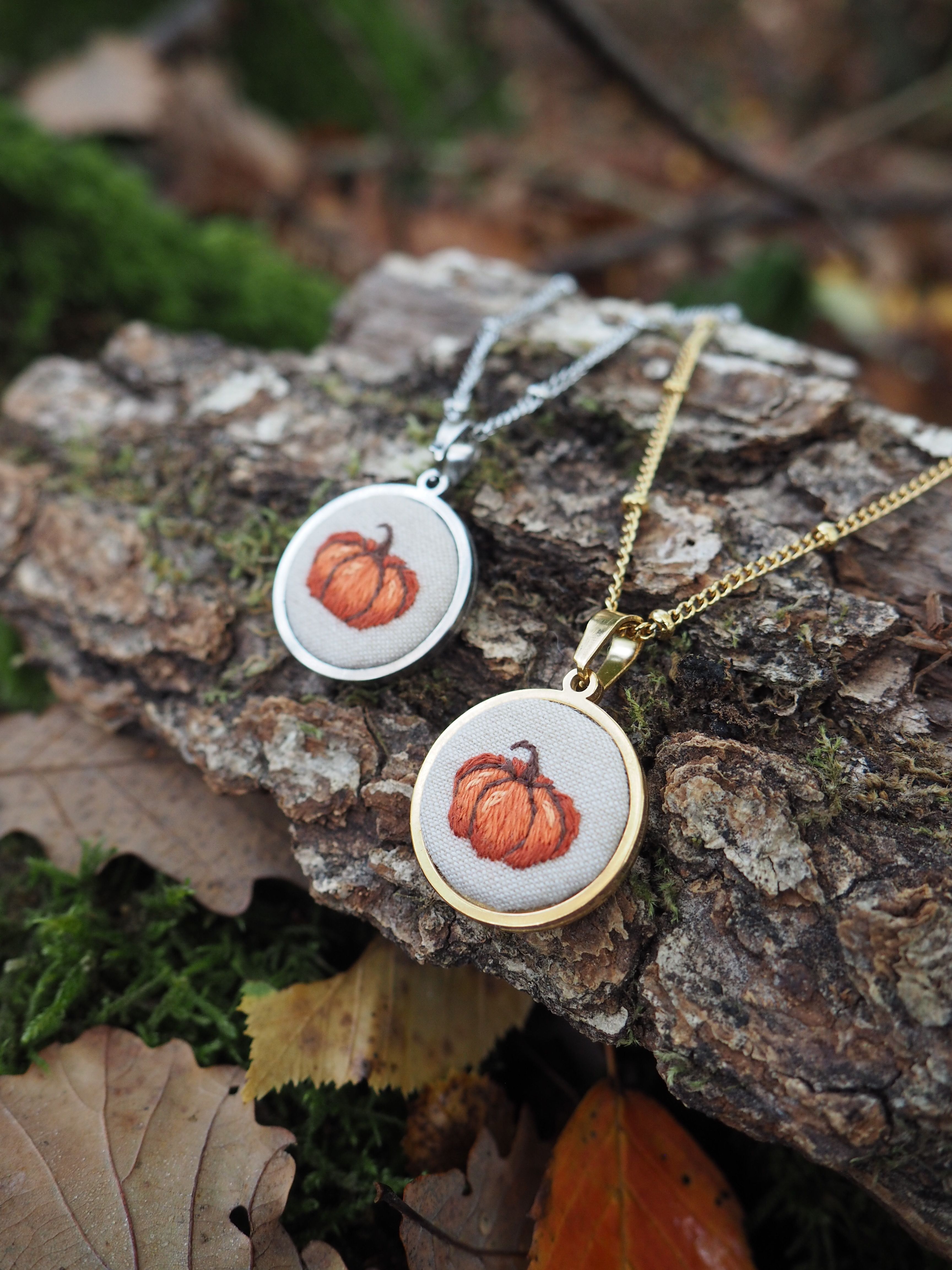 Cette image présente deux colliers pendentifs disposés sur un morceau d'écorce dans un environnement naturel automnal.  **Les bijoux :**
- Deux pendentifs circulaires identiques avec des montures dorées
- Chacun contient une broderie délicate représentant une citrouille orange
- Un collier a une chaîne argentée, l'autre une chaîne dorée
- Le travail de broderie est fin et détaillé, donnant un aspect artisanal **Le décor :**
- Les pendentifs reposent sur un morceau d'écorce texturée
- L'arrière-plan présente des feuilles mortes dans les tons bruns et dorés
- On aperçoit de la mousse verte qui ajoute une touche de couleur naturelle
- L'ensemble évoque parfaitement l'atmosphère automnale **L'ambiance :**
Cette mise en scène photographique met en valeur ces bijoux à thème saisonnier, parfaits pour célébrer l'automne ou Halloween. La présentation sur des éléments naturels renforce le côté authentique et fait-main de ces accessoires.