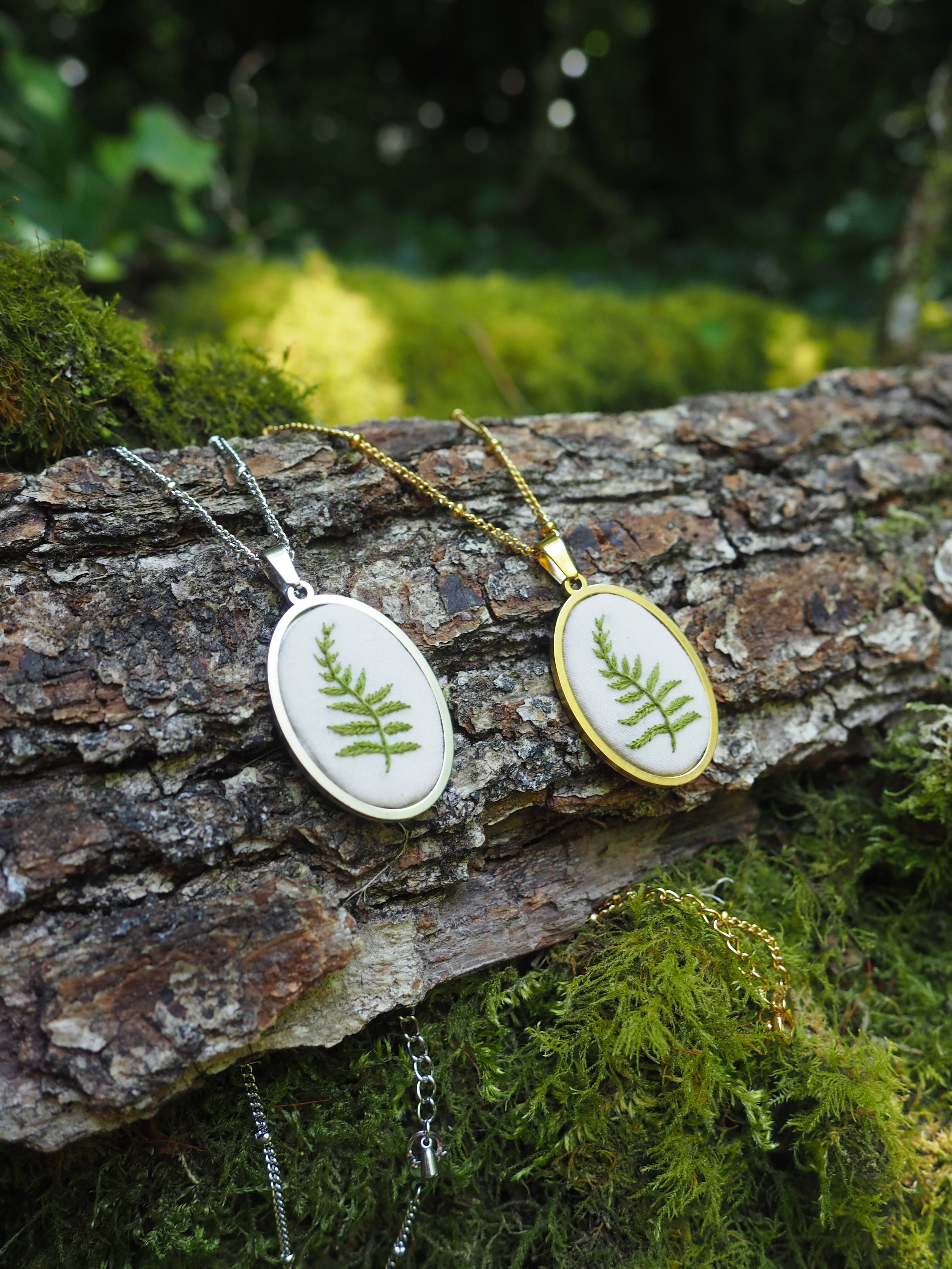 L'image montre des bijoux artisanaux délicats posés sur un morceau d'écorce d'arbre couvert de mousse. Ce sont des pendentifs qui semblent contenir de véritables feuilles de fougère préservées à l'intérieur de petits cadres blancs et dorés. Les pendentifs sont attachés à des chaînes argentées et créent un beau contraste avec le fond naturel texturé de l'écorce et la mousse verte. La composition est très naturelle et évoque un style botanique élégant, mêlant la nature et l'artisanat.
