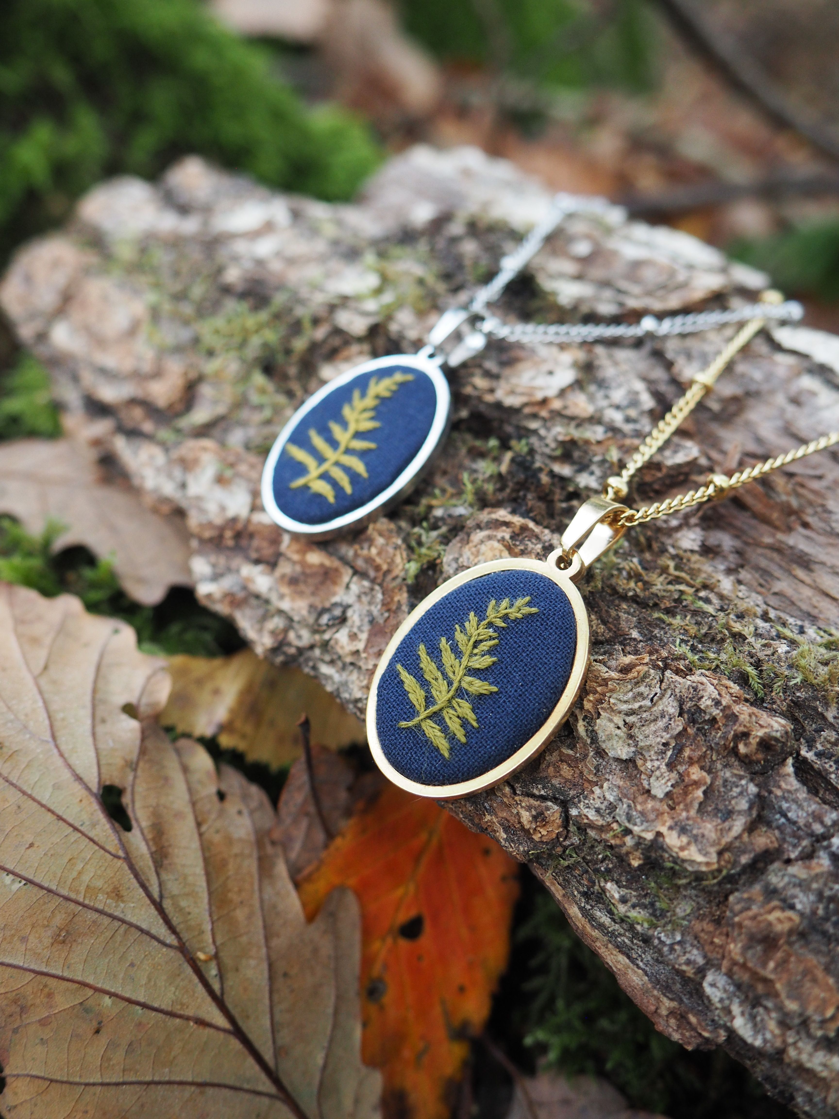 Cette image présente deux bijoux artisanaux élégants posés sur un morceau d'écorce dans un environnement naturel automnal. **Description des bijoux :**
- Deux pendentifs de forme ovale avec des montures dorées
- Fond bleu marine profond sur lequel sont incrustées de délicates fougères dorées
- Les chaînes sont fines et dorées, donnant un aspect raffiné
- Le style évoque la bijouterie botanique ou la résine époxy avec inclusions végétales **Environnement :**
- Les pendentifs reposent sur un morceau d'écorce rugueuse et texturée
- Autour, on aperçoit des feuilles d'automne dans des tons chauds (orange, brun)
- L'arrière-plan est naturellement flouté avec de la verdure
- L'éclairage naturel met en valeur les détails dorés des bijoux **Impression générale :**
Ces pièces semblent être des créations artisanales inspirées de la nature, parfaites pour quelqu'un appréciant les bijoux botaniques. La mise en scène dans cet environnement forestier souligne harmonieusement le thème naturel des pendentifs.
