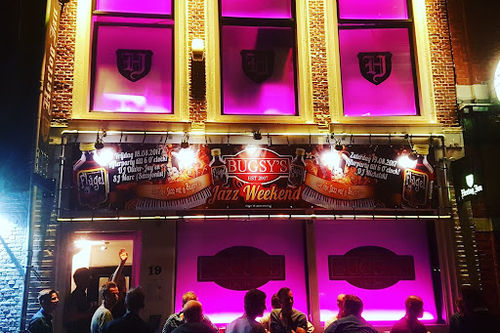 Bugsy's live music club exterior at night with illuminated pink windows, stage areas visible, and crowd gathered at entrance.