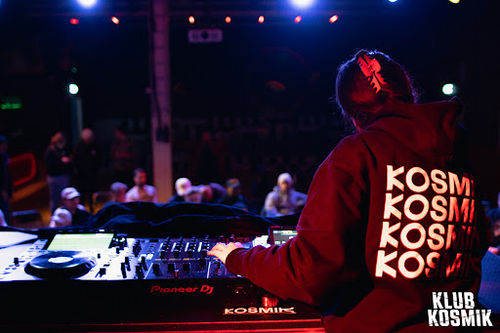 DJ performing at KOSMIK bar with turntables, red lighting, and crowd dancing on dance floor.