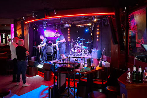 Café Miles live music venue with illuminated stage, band performing with drums and instruments, red neon lighting, and audience seating area.