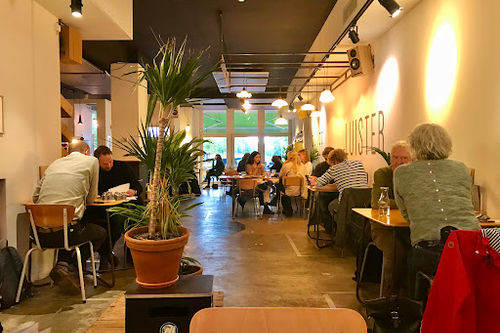 Het Muzieklokaal bar interior with stage lighting, potted plants, wooden tables, and seated patrons in warm ambient lighting.