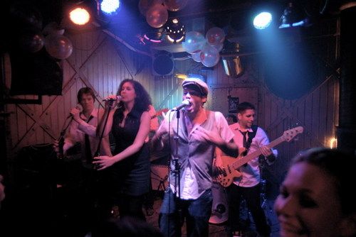 Live band performing at Cafe Stiels with stage lighting, PA system, and audience in intimate venue setting.