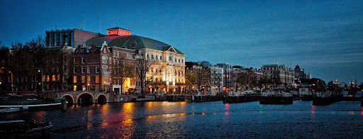Royal Theater Carré illuminated at dusk along Amsterdam's canal, historic concert hall architecture reflected in water.