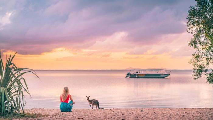 Woman kneelin on a beach besides a small kangaroo at sunrise