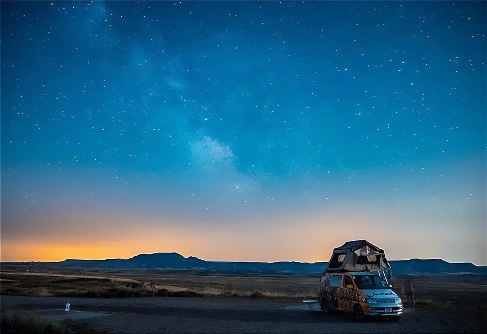 Campervan with rooftop tent sits in a vast ladscape under a starry night sky, a glow on the horizon