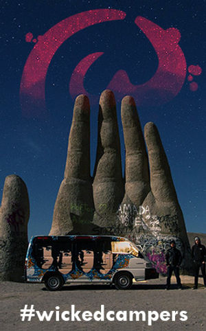 Men standing beside griffiti art campervan, in front of a giant statue of a hand in the desert under a starry sky, text at the bottom reads '#wickedcampers'