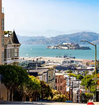 Looking down a hill in San Francisco city, California with buildings, trees, cars, changes in elevation, and water.