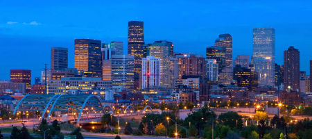 Denver, Colorado state city skyline with buildings lit up by warm colored lights and a bridge outlined by silver arches.