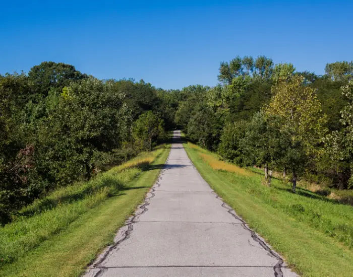 Volksweg Trail in Pella, Iowa with a straight cement road leading down the middle between groves of green trees and grass on a sunny game.