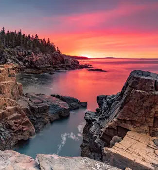 Landscape scene of Arcadia National Park in Maine at sunset dusk with red warm hues and large body of water between rocks and forestry.