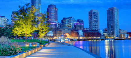 Boston, Massachusetts waterfront at night with a large pathway, trees, and the city skyline reflecting on the water.