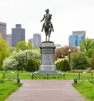 Statue in Boston Common Park, Massachusetts of a man riding a horse on a pedestal with buildings behind trimmed shrubbery and a variety of trees.