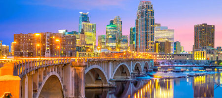 Downtown Minneapolis in Minnesota at dawn with bright yellow light reflecting off buildings and water under a bridge.