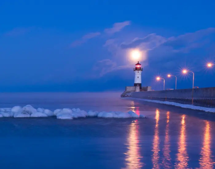 Lighthouse in Duluth Minnesota at night with sea foam and snow flowing towards the shoreline and a long walking path to the white and red lighthouse.