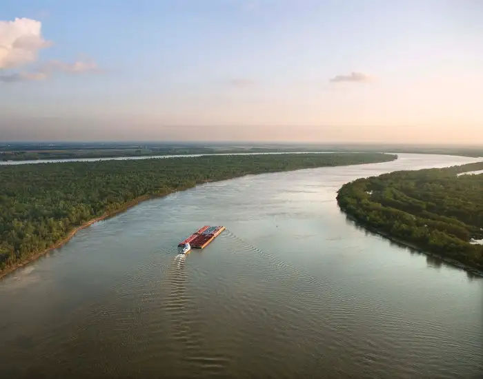 Red boat going down the Mississippi River with calm waters surrounded by thick natural green forest trees.