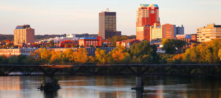 Manchester, New Hampshire city skyline at dusk with warm sunlight bouncing off tall buildings behind a waterfront.