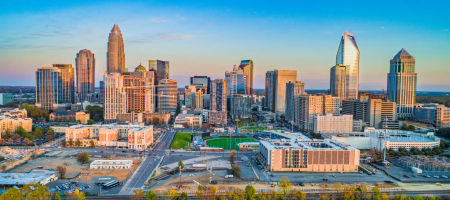 North Carolina city skyline during sunrise with warm orange hues of light beaming on numerous buildings with a green stadium in the center.